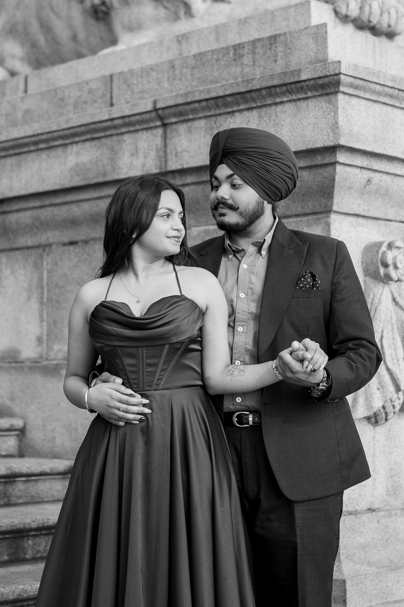 A romantic black and white portrait of a couple in formal attire posing against a stone wall outdoors.