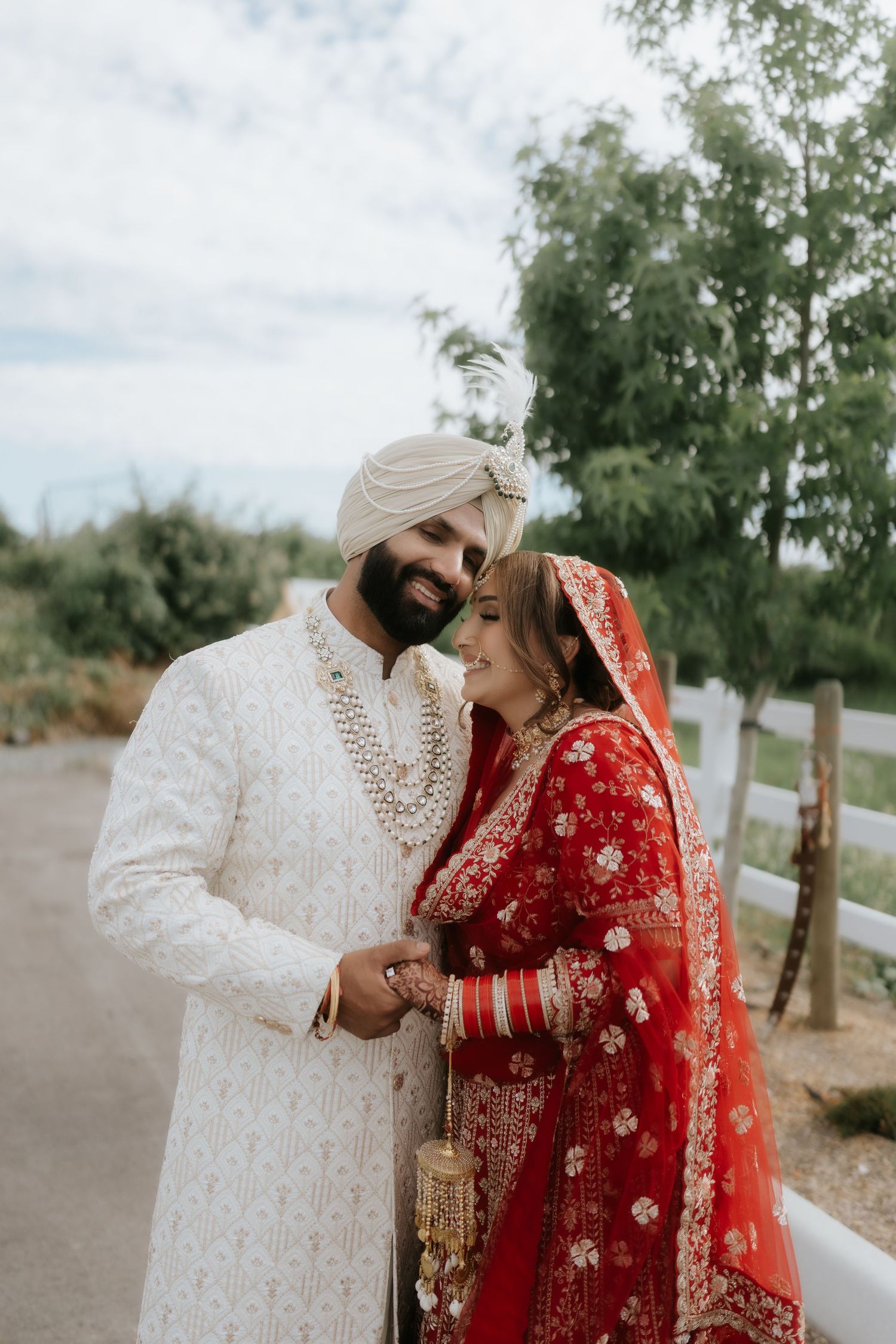 Sikh bride in red lehenga and groom in white sherwani smiling together outdoors on their wedding day.