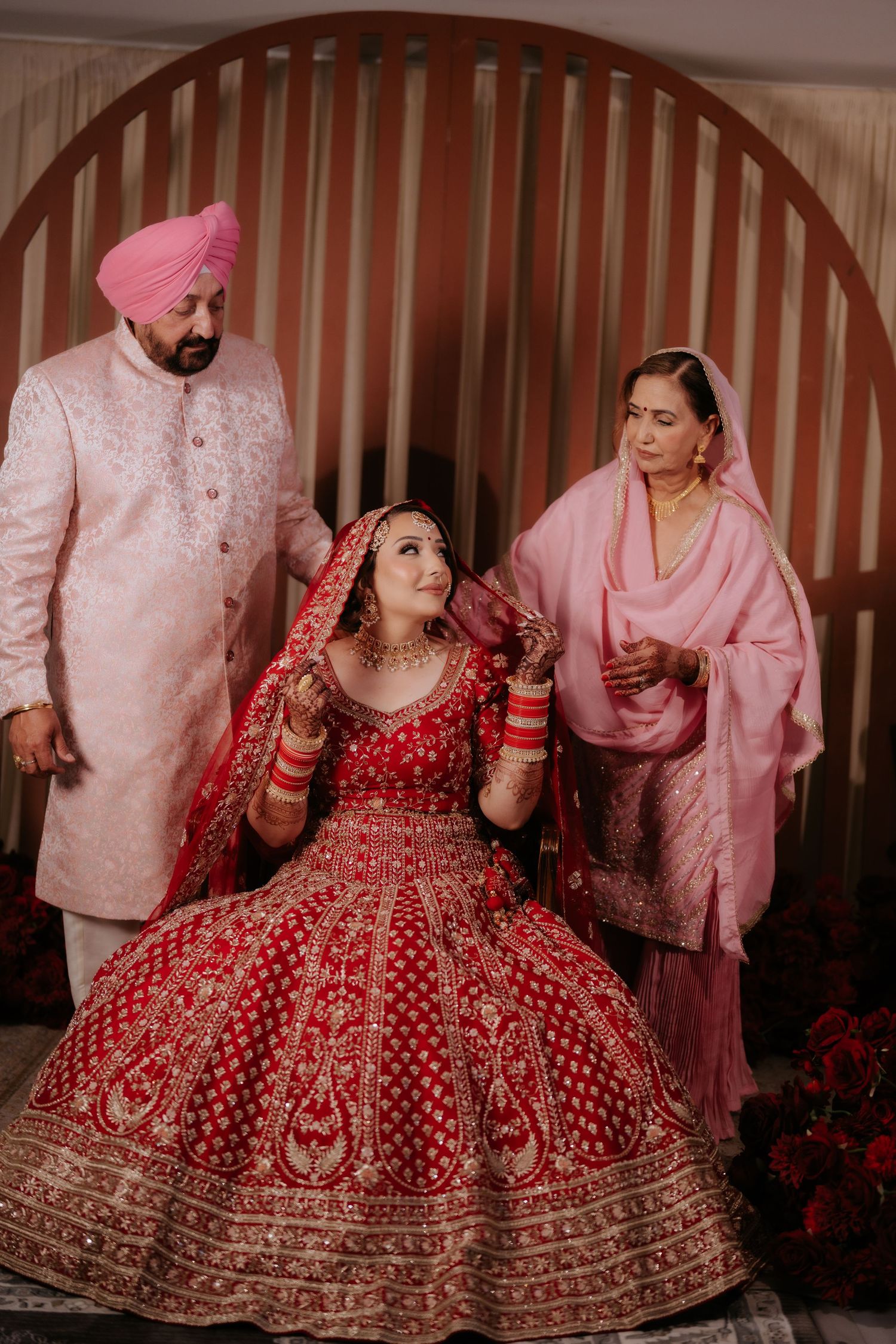 Bride in red bridal lehenga with gold embroidery seated, flanked by parents in traditional Punjabi attire.