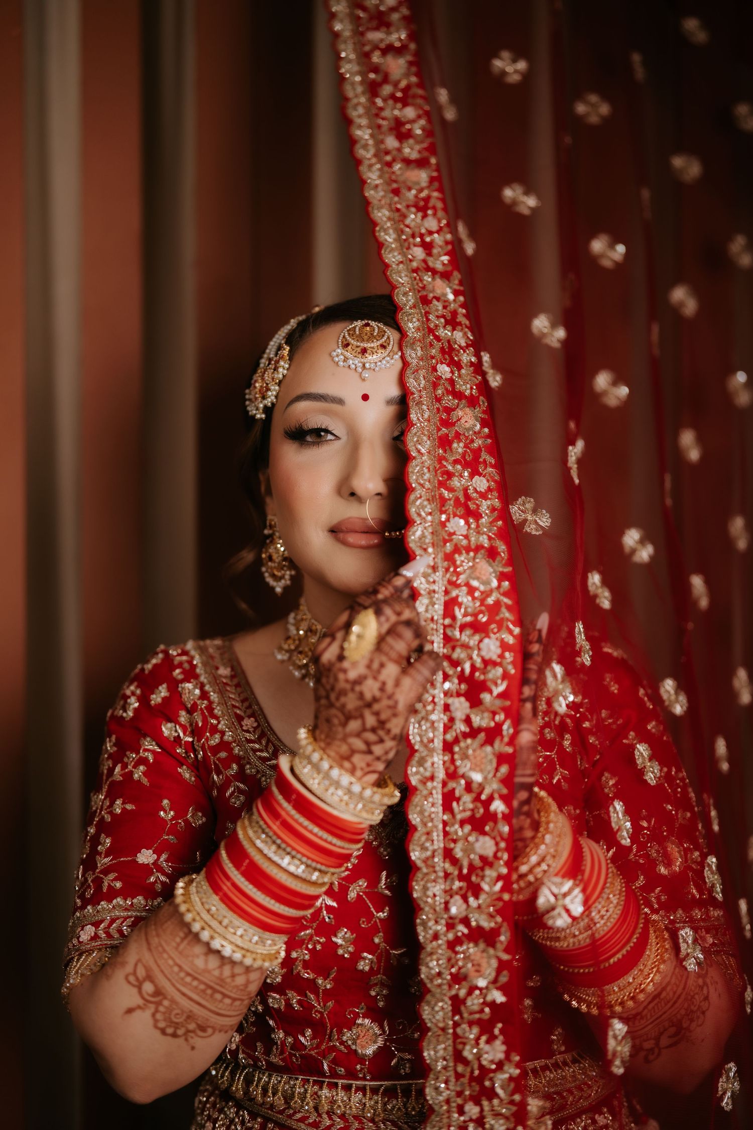 Indian bride in red lehenga with gold embroidery holds dupatta, adorned with traditional jewelry and mehndi.