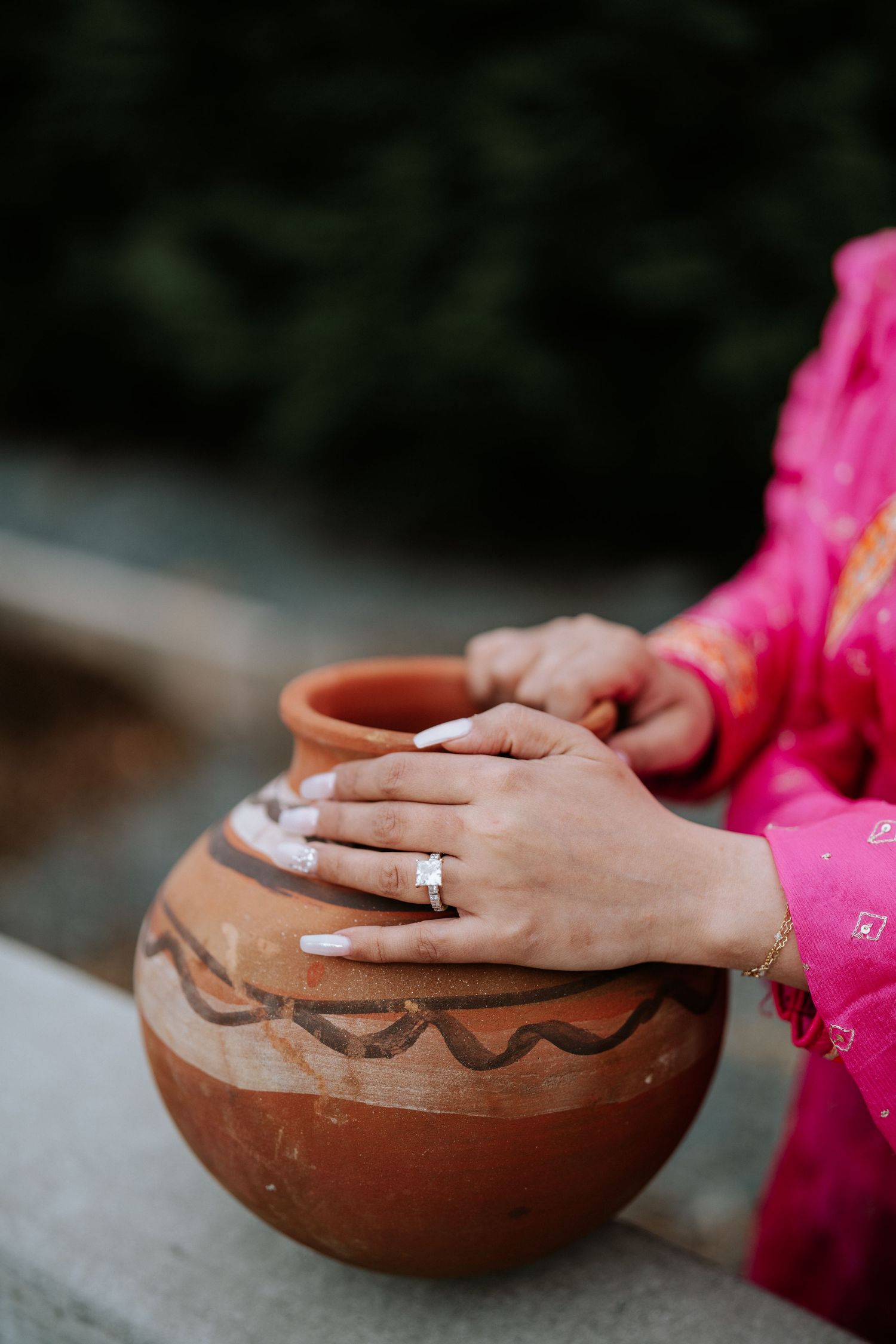 Woman in pink sari holding a traditional terracotta clay pot with decorative patterns, wearing white nail polish and gold ring.