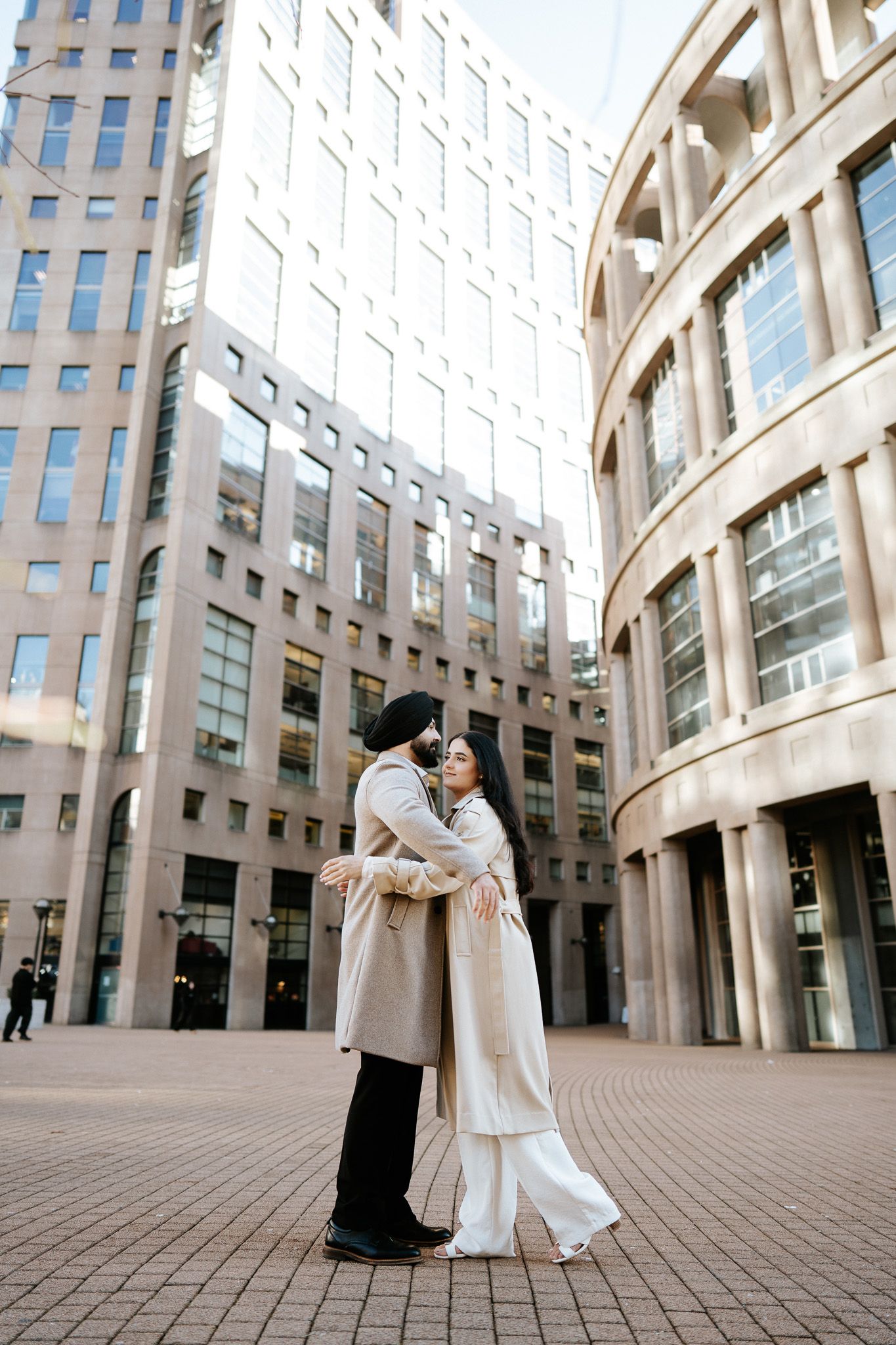 Two people in light colored coats embrace in front of modern glass office buildings in an urban plaza.