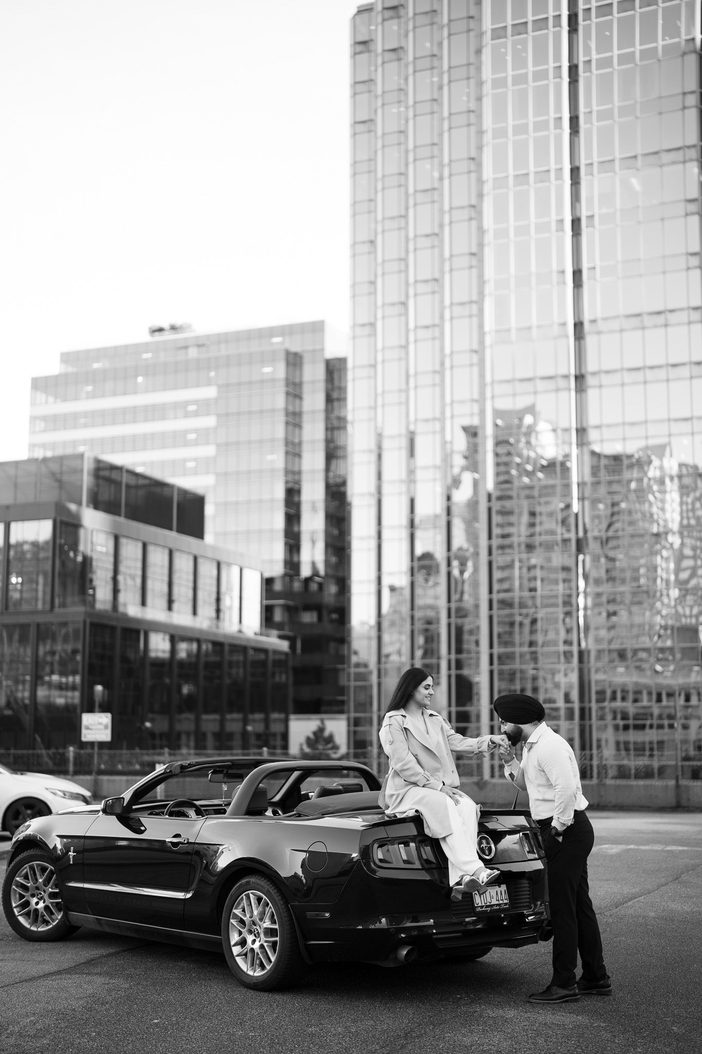 A couple chats while sitting on a black Mustang convertible in front of modern glass office buildings in black and white.