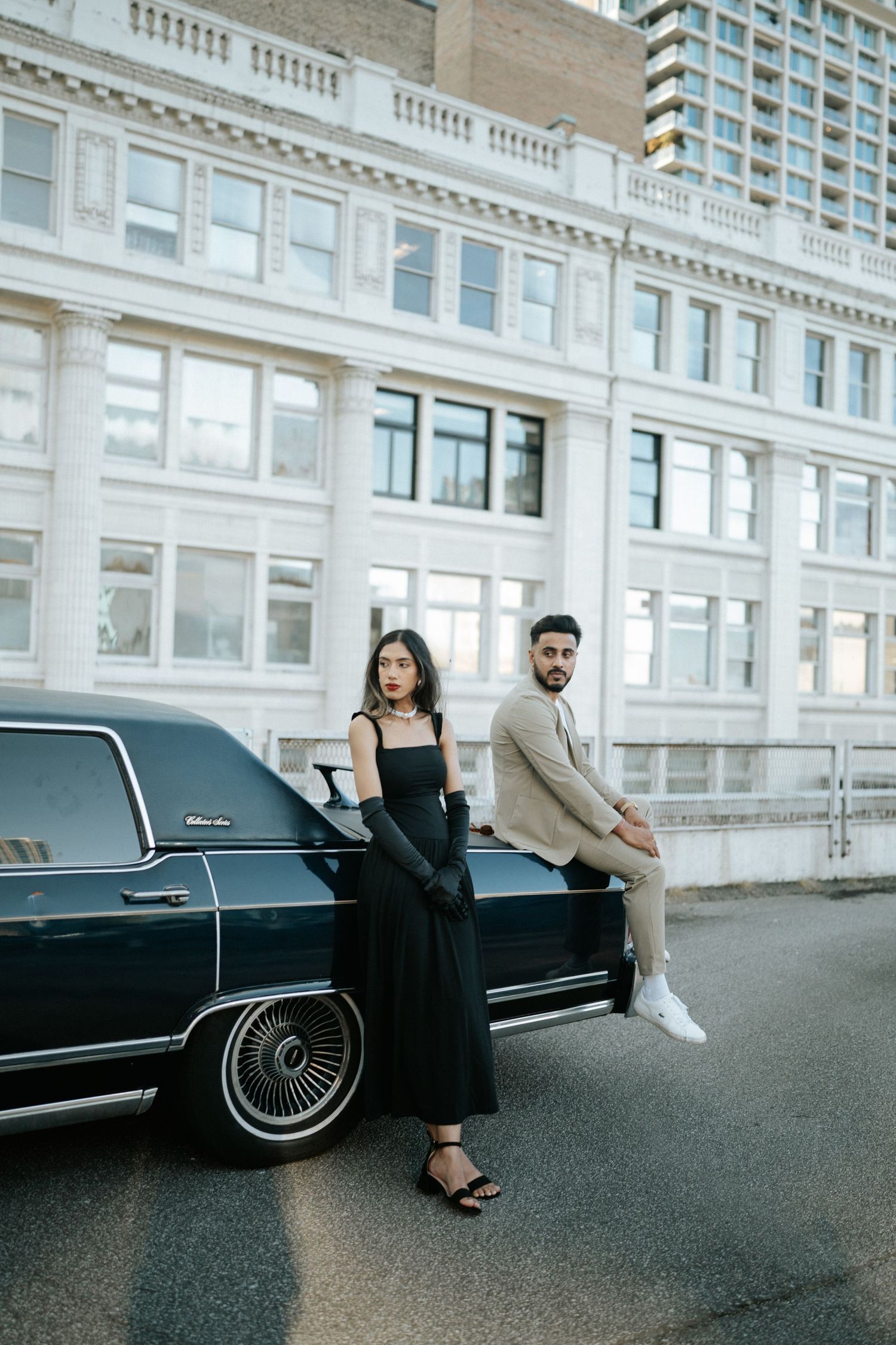 Couple posing elegantly next to a vintage black limousine on a city street with white classical architecture.