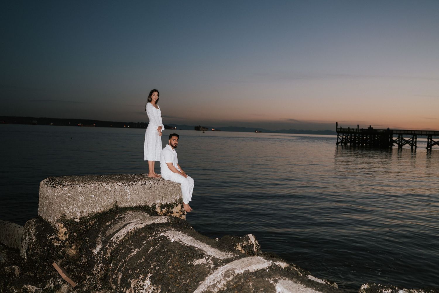 Scenic coastal sunset photo shoot with white dress against dramatic ocean and rocky shoreline backdrop.