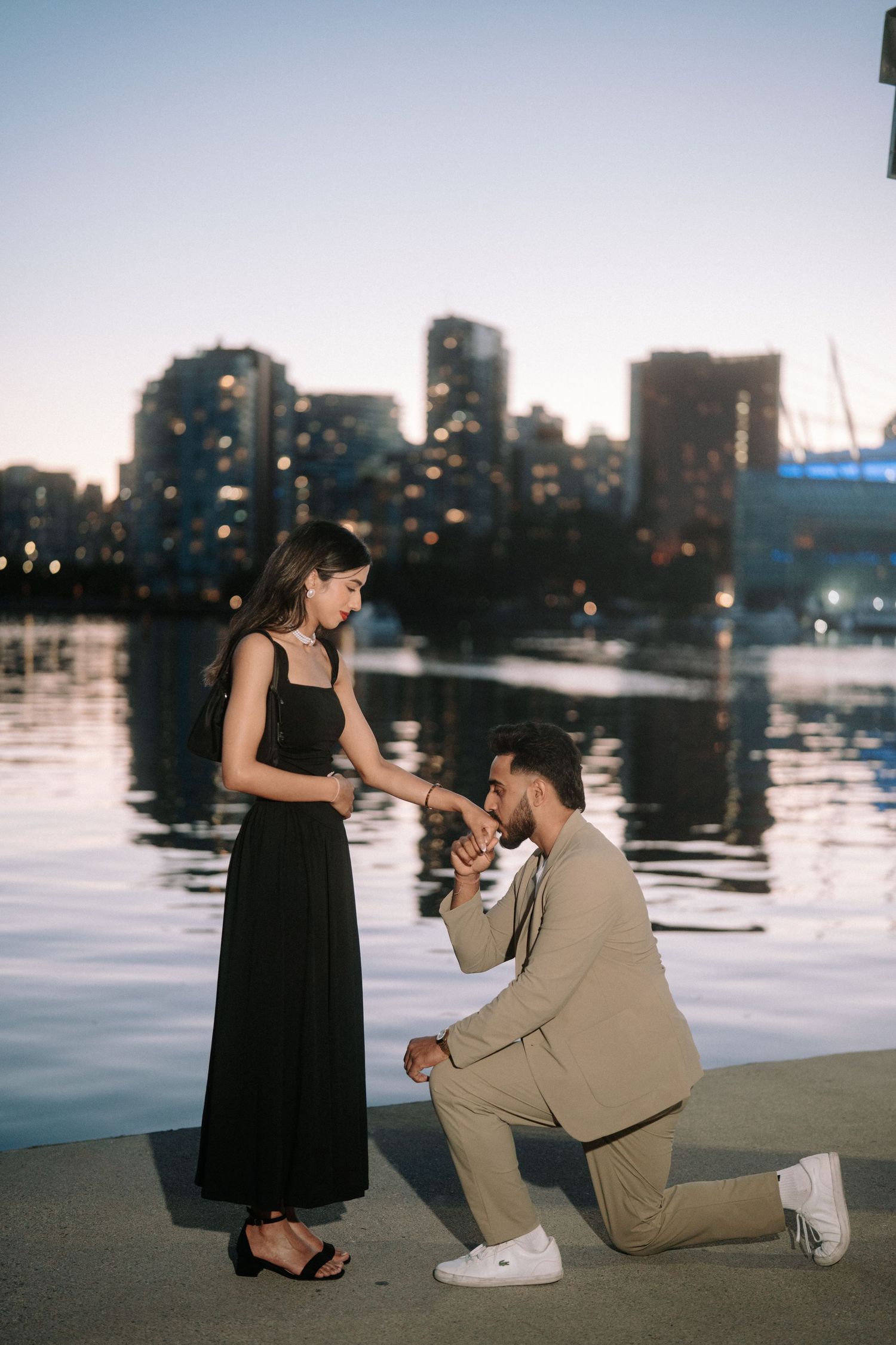 A couple shares a romantic proposal moment by the waterfront at sunset with city skyline in the background.