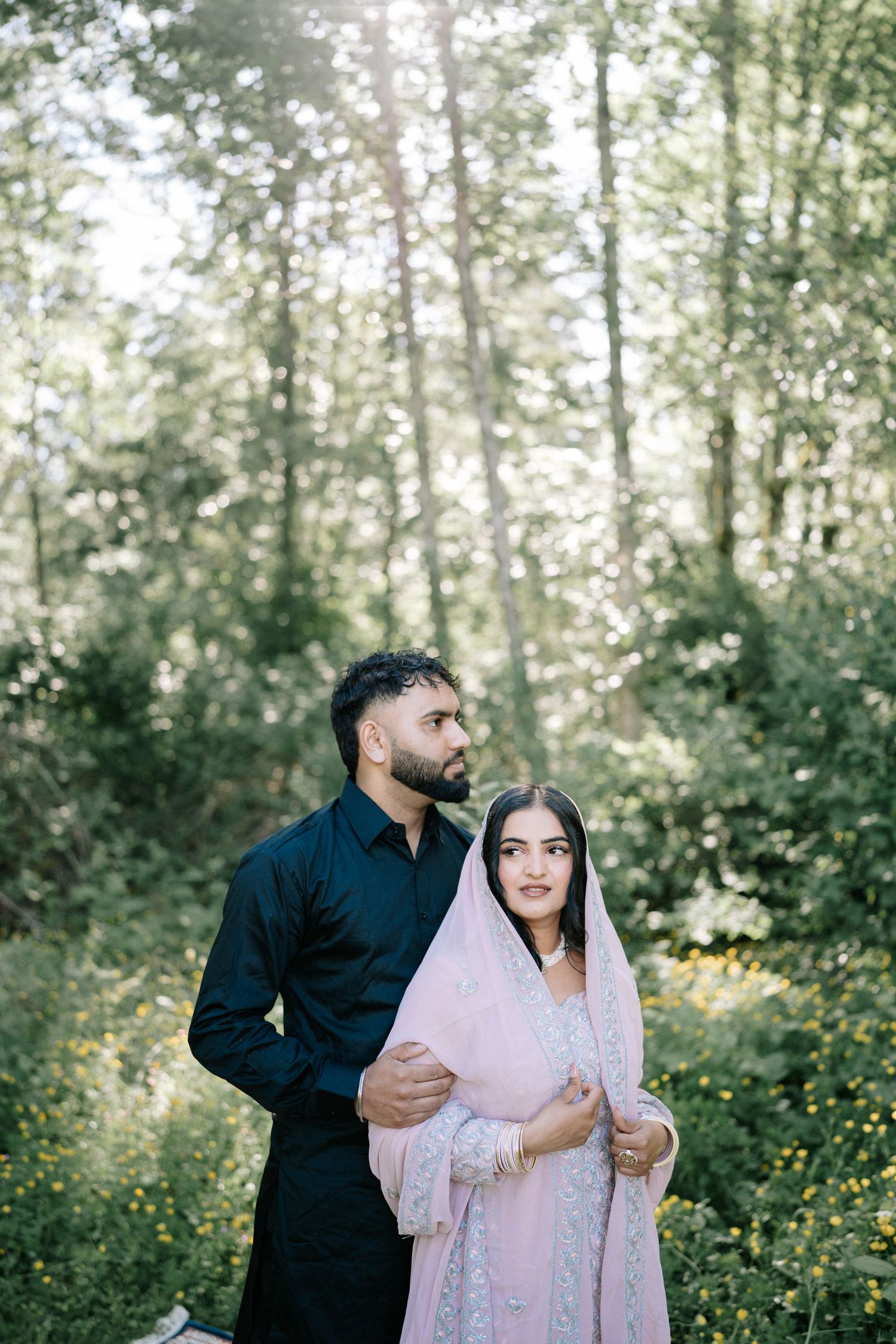 Couple in traditional attire pose together in a sunlit forest setting.