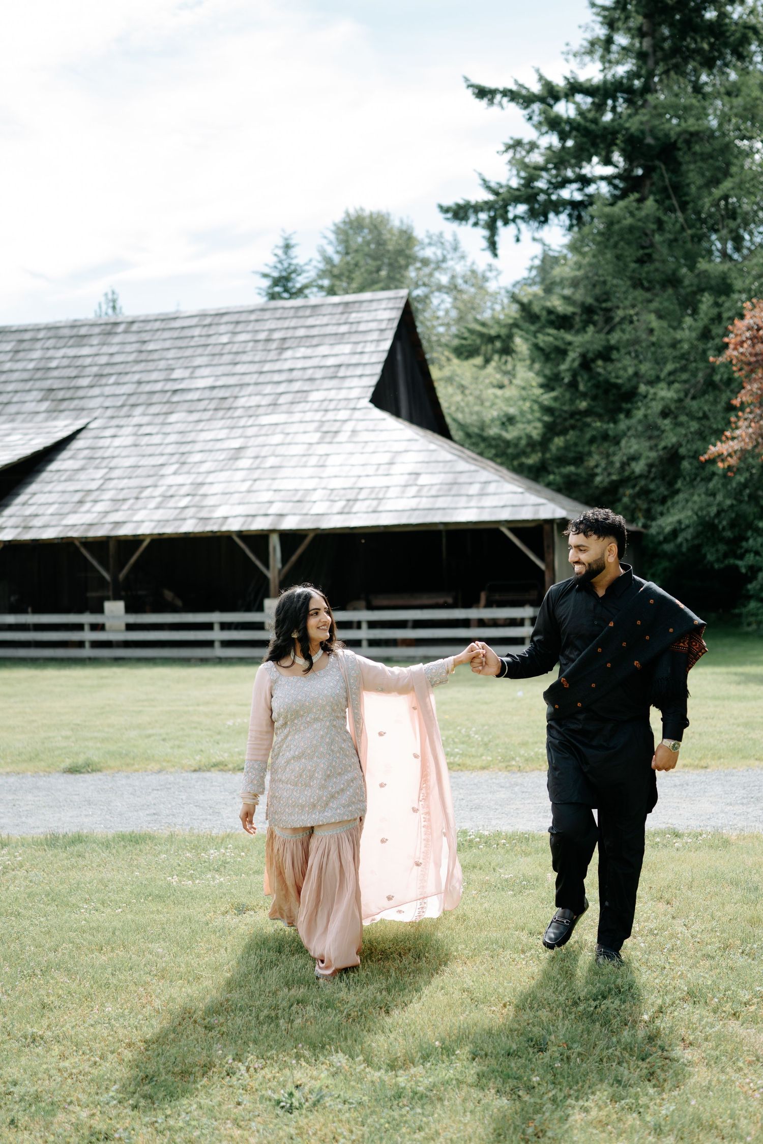 A couple in formal attire walks hand in hand through a garden with rustic buildings in the background.