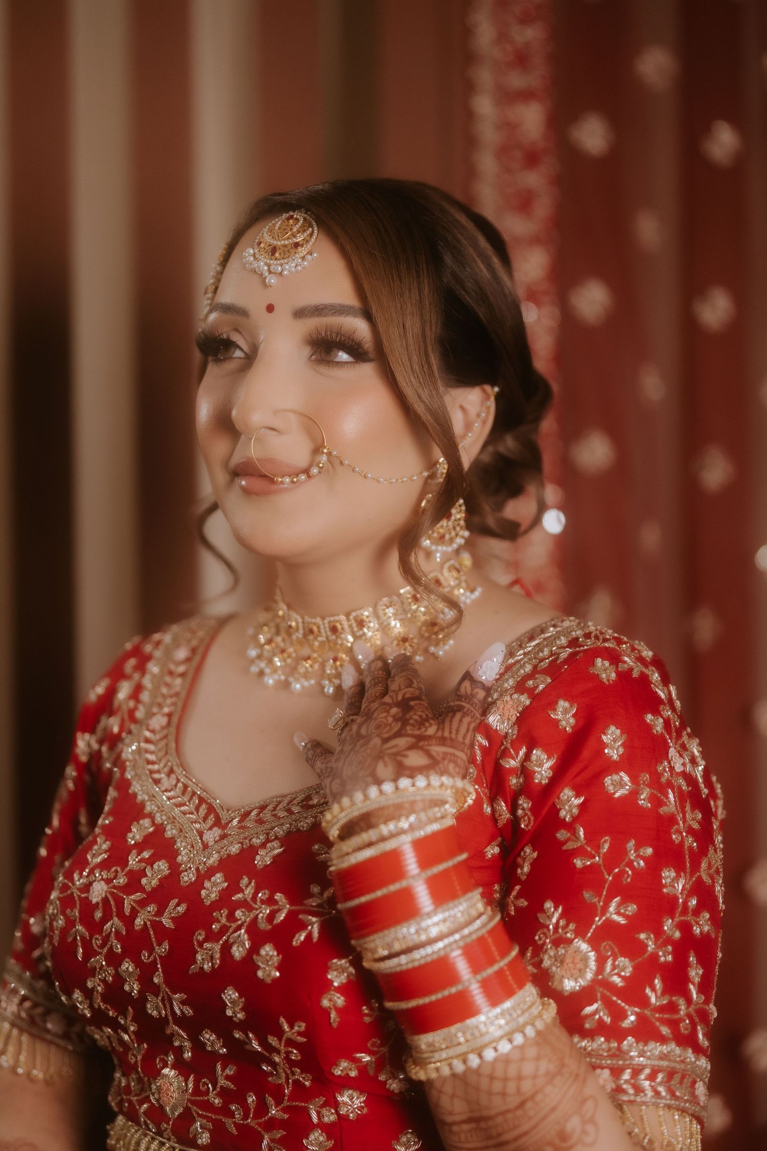 Indian bride in red lehenga with gold embroidery, wearing traditional bridal jewelry including maang tikka and nath.