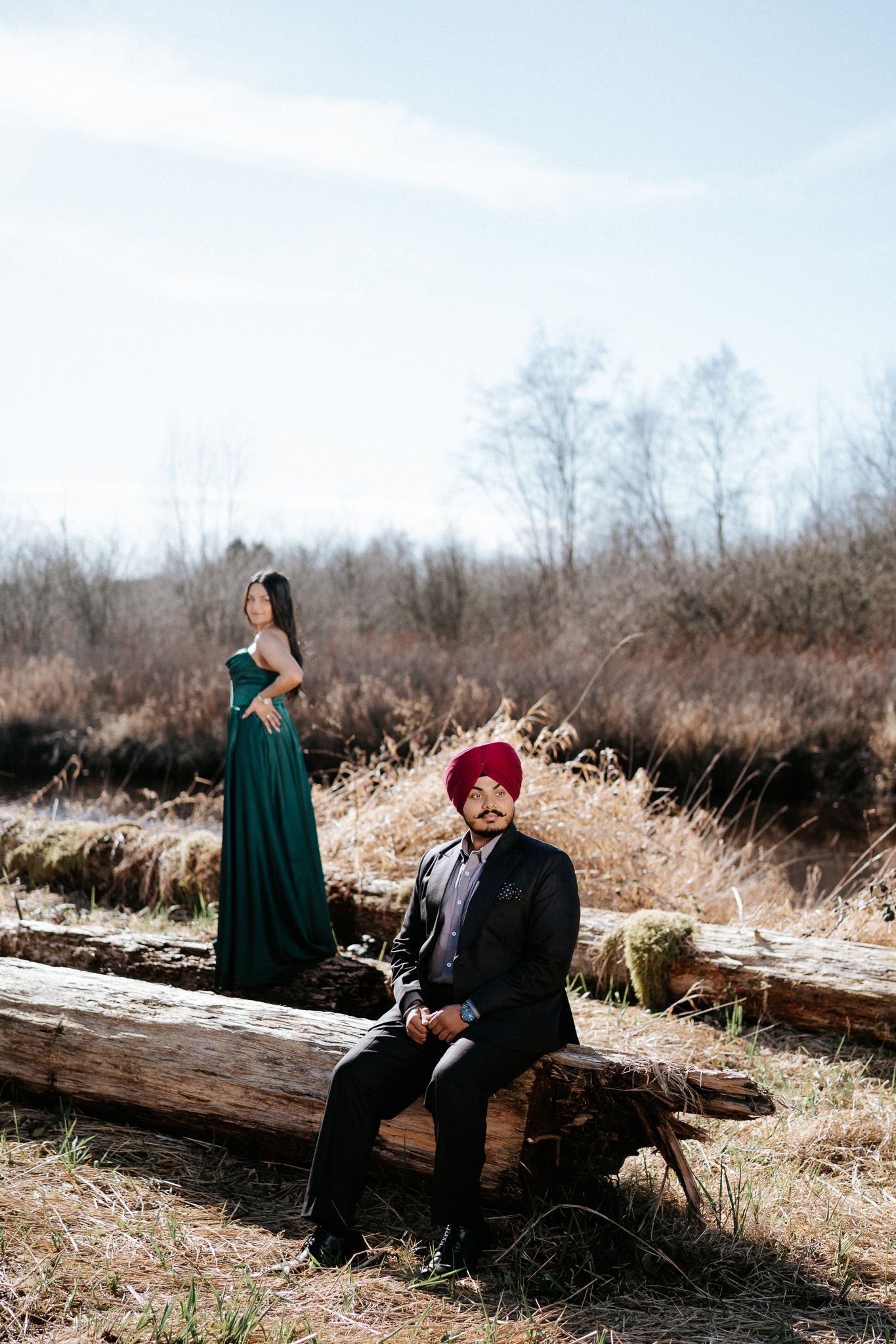 A couple sits together on a fallen log in a winter field with dried grass and bare trees in the background.