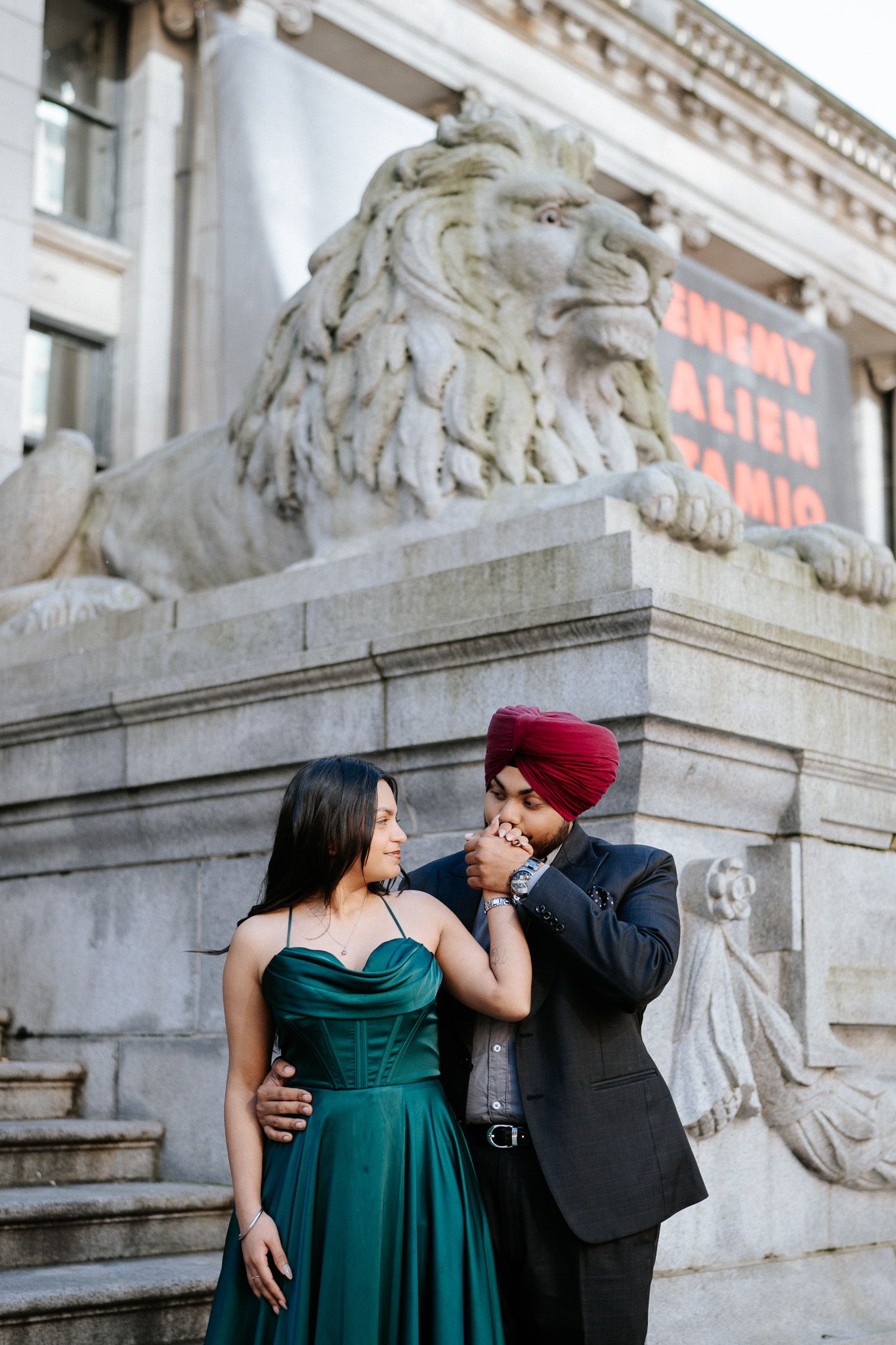 A couple poses for romantic engagement photos in front of an ornate stone building with decorative sculptures.