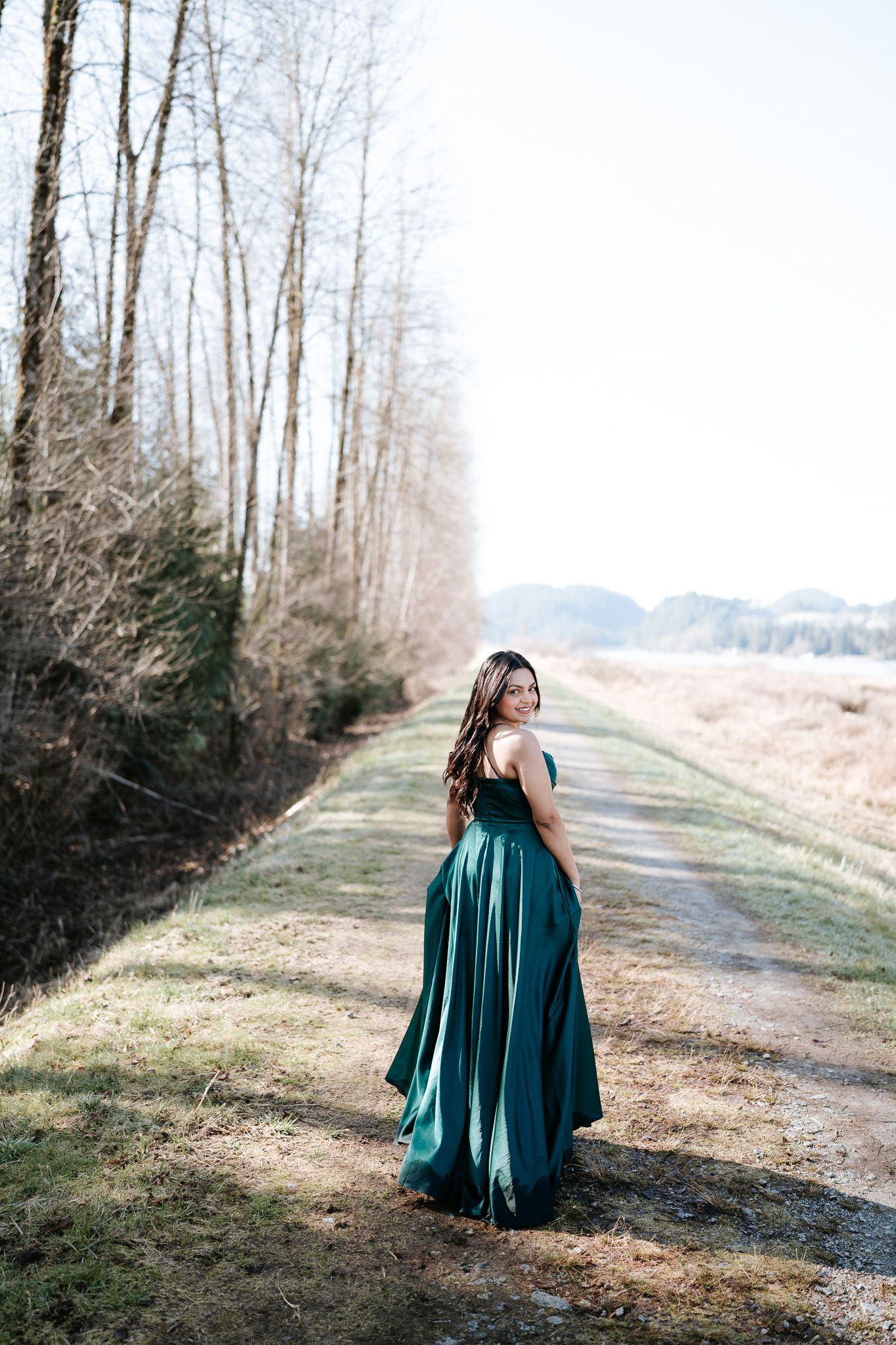 A figure in an elegant emerald green gown stands on a serene forest path during a photo session.