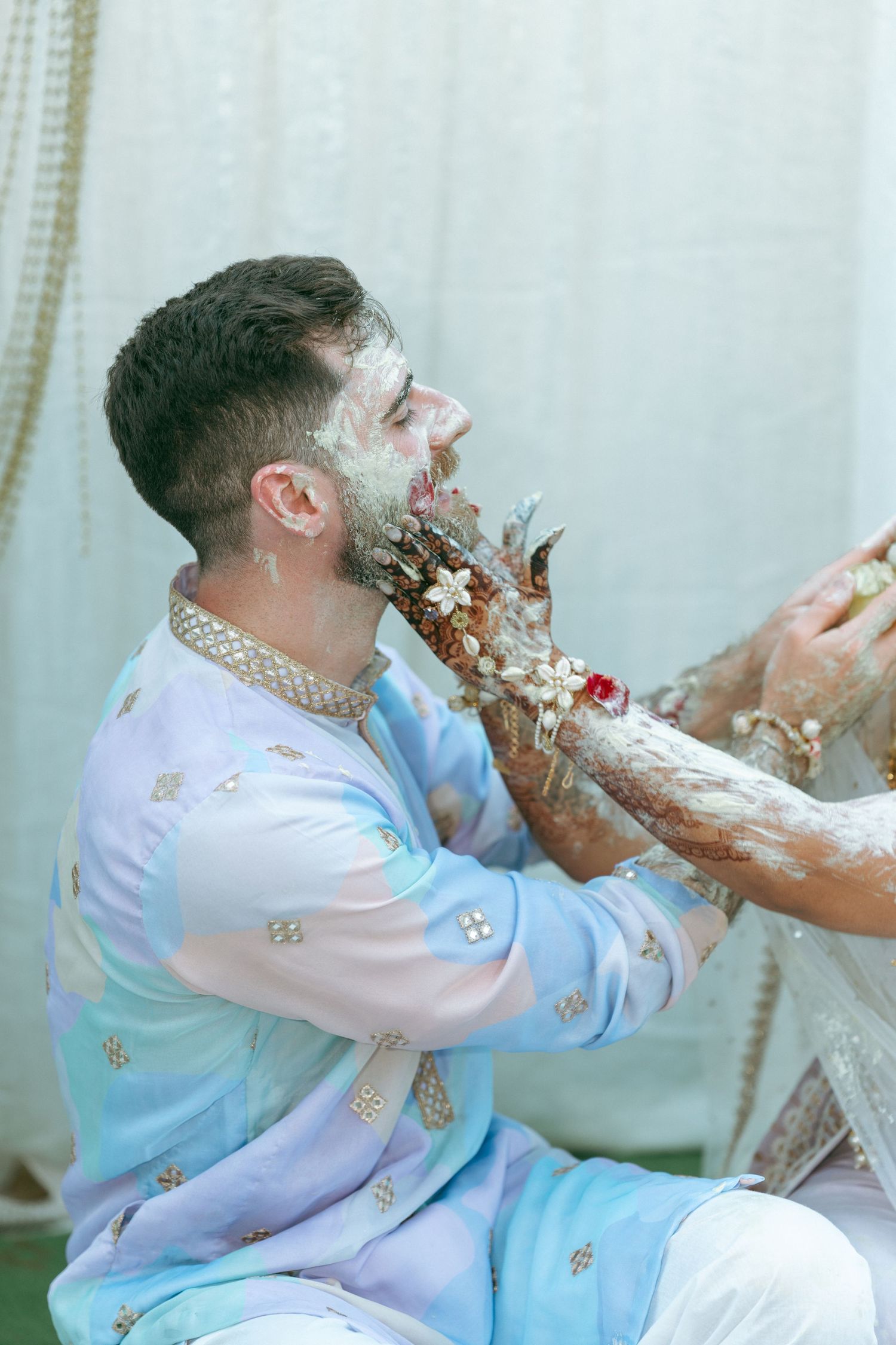 Groom in blue kurta gets haldi applied by mehndi-adorned hands during Indian wedding ceremony.