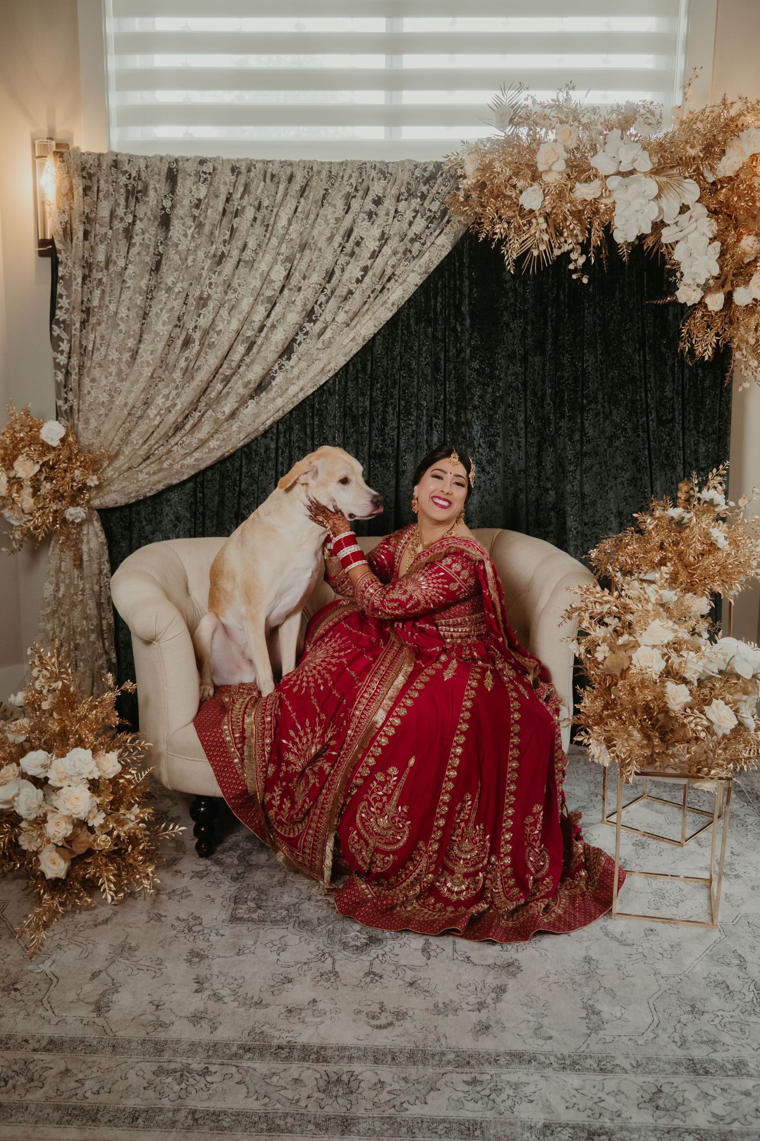 Bride in red and gold lehenga sits with white dog on cream sofa, surrounded by dried floral arrangements.