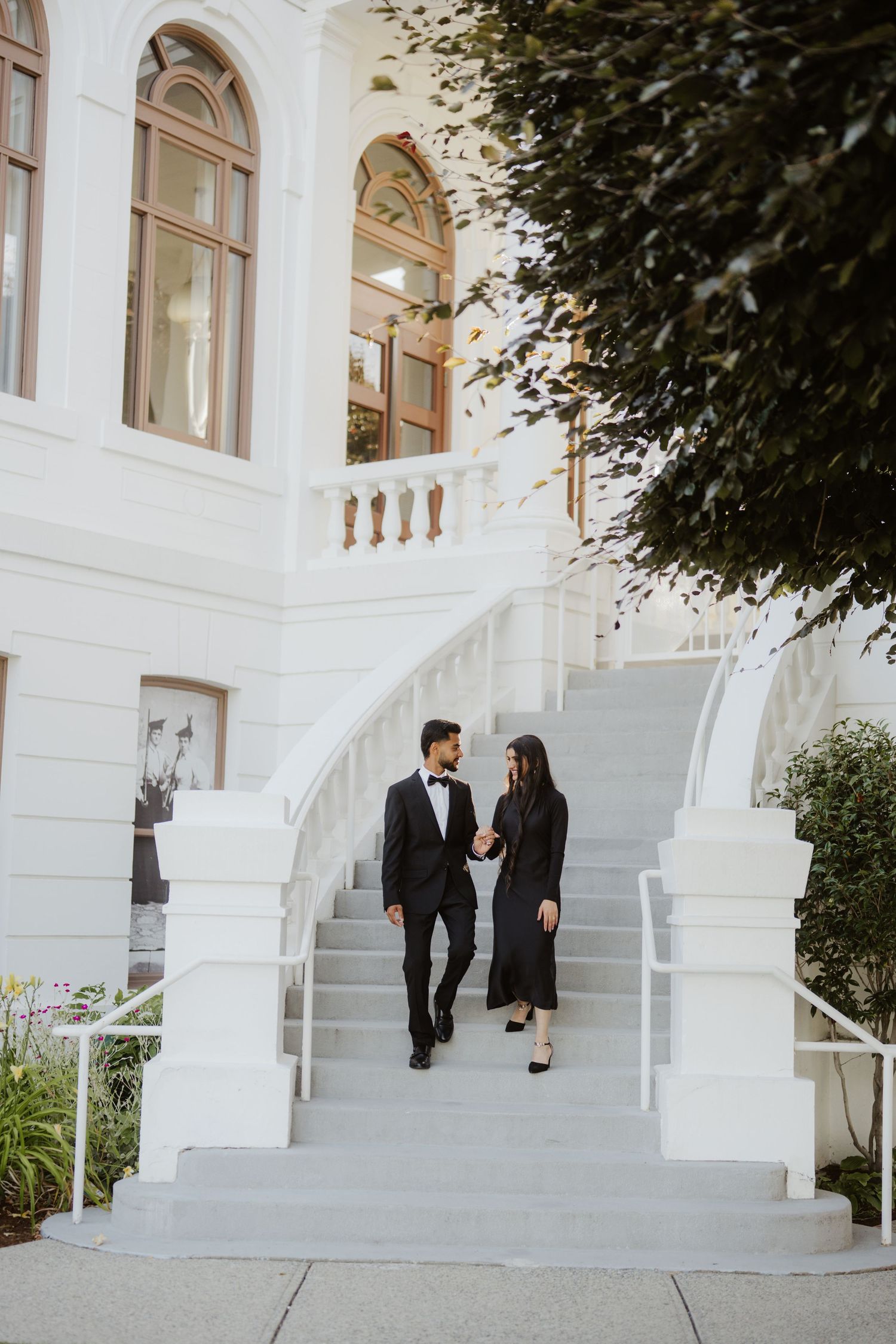 Elegant couple in formal black attire walking down white grand staircase of historic building.