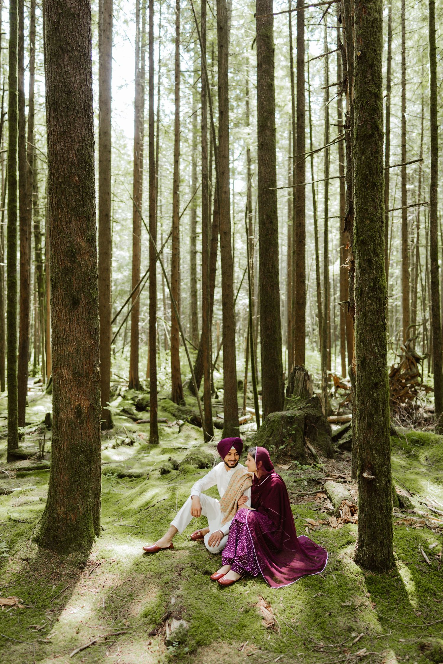 Sikh couple in traditional attire sitting on mossy forest floor surrounded by tall trees.
