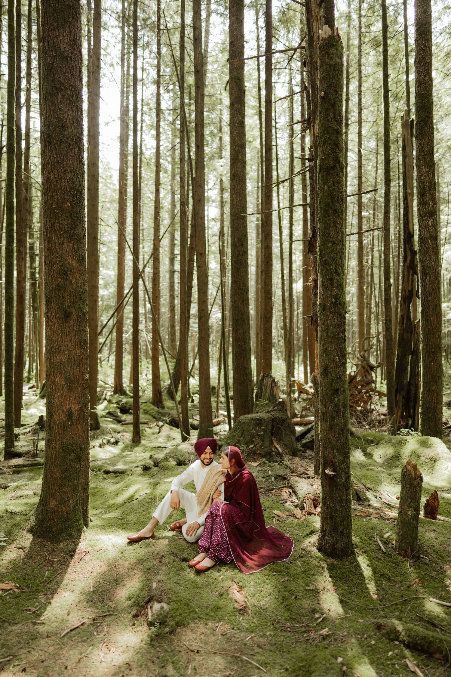 Sikh couple in traditional attire sitting together on mossy forest floor surrounded by tall trees.