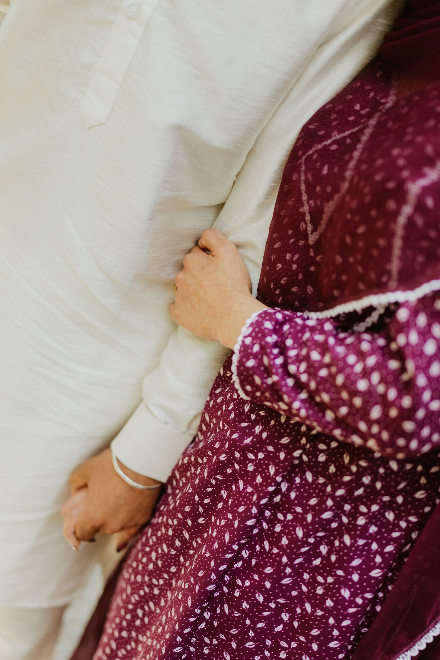 Close-up of woman in maroon embroidered salwar holding man's white kurta sleeve, gold bangle visible.