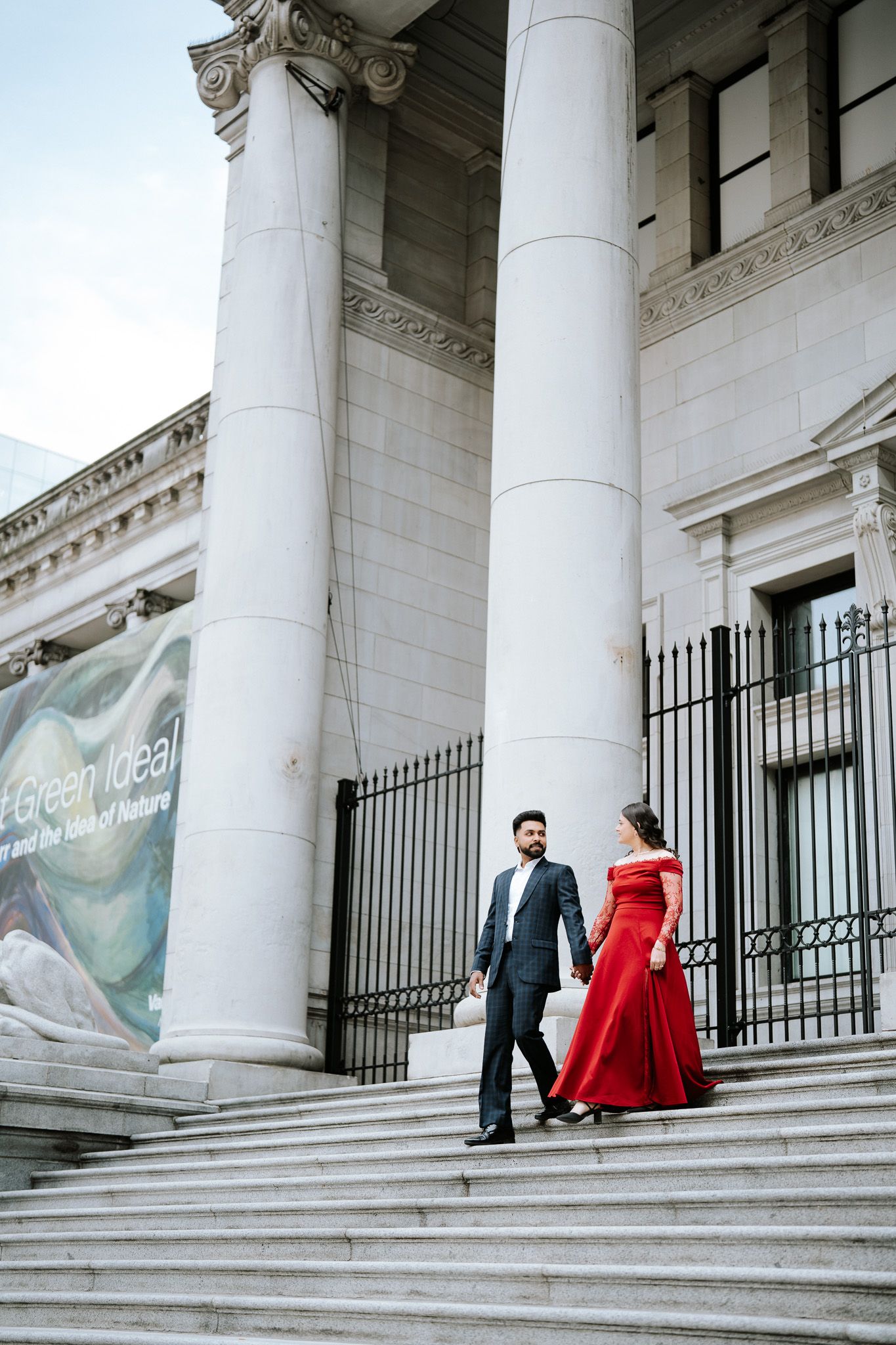 Couple in formal attire descending grand stone steps of a neoclassical building, woman in red gown, man in black suit.