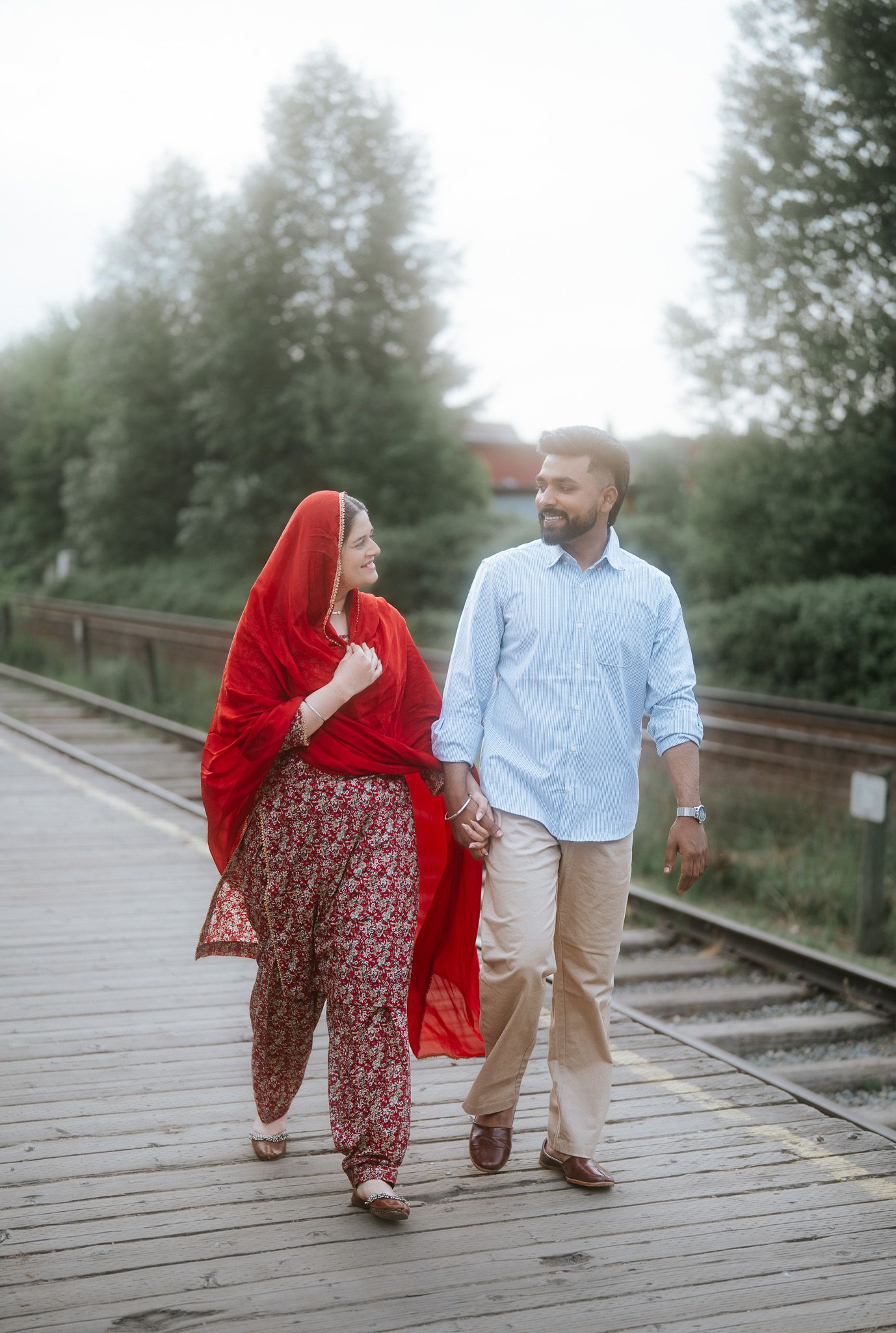 Couple walking hand in hand on wooden railway platform, woman in red dupatta and floral salwar suit.