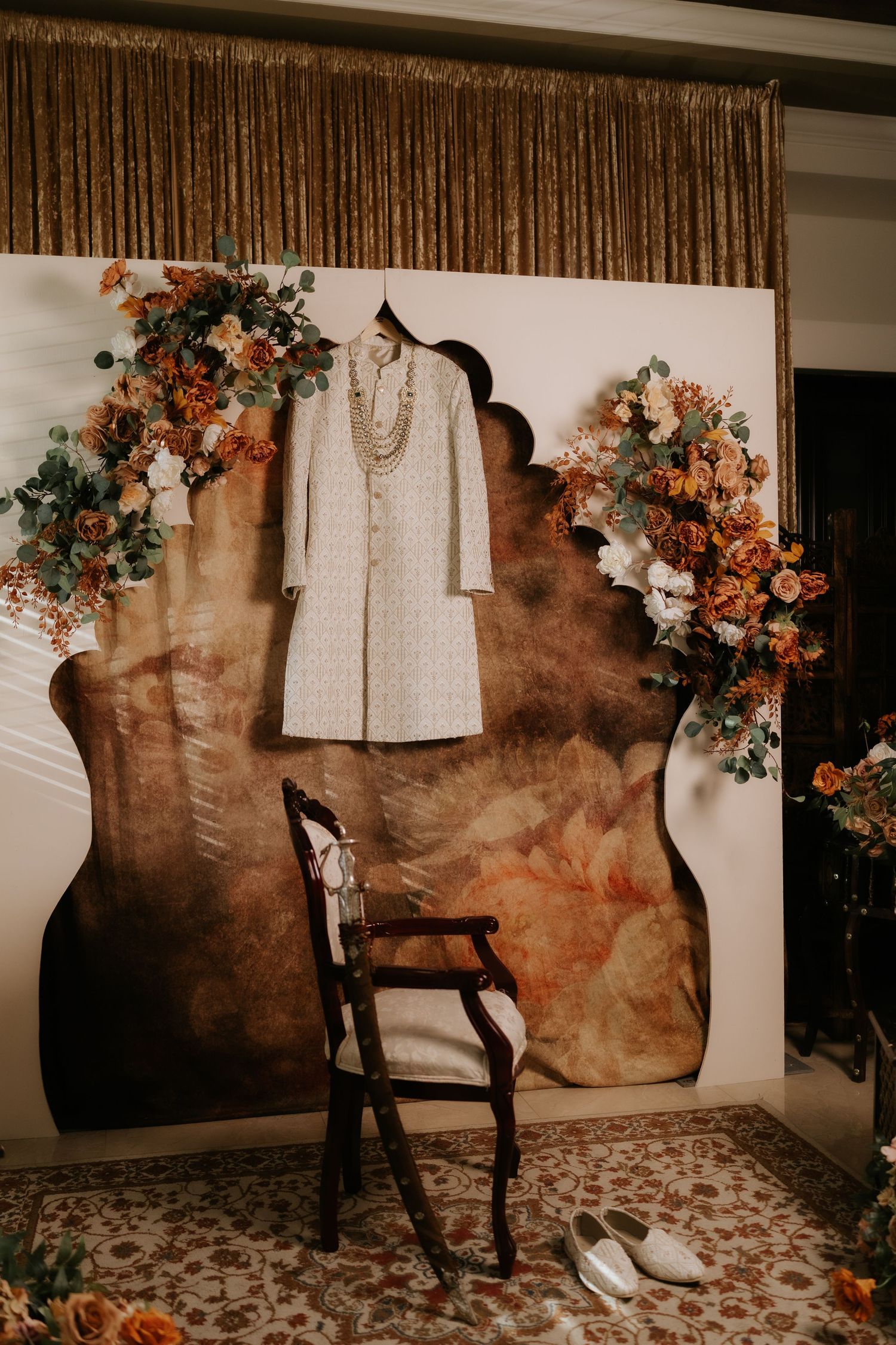 Elegant groom's sherwani displayed on a decorative backdrop with floral arrangements and pearl necklace at an Indian wedding.