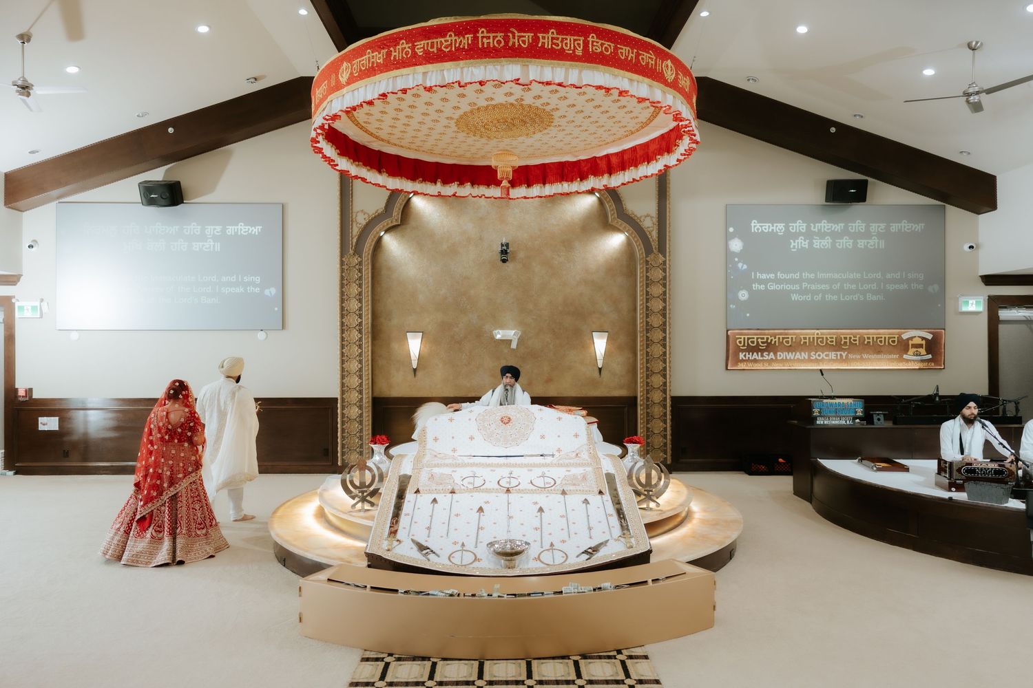Sikh Gurdwara interior with ornate Palki Sahib, red canopy, and worshippers in traditional attire during a religious ceremony.
