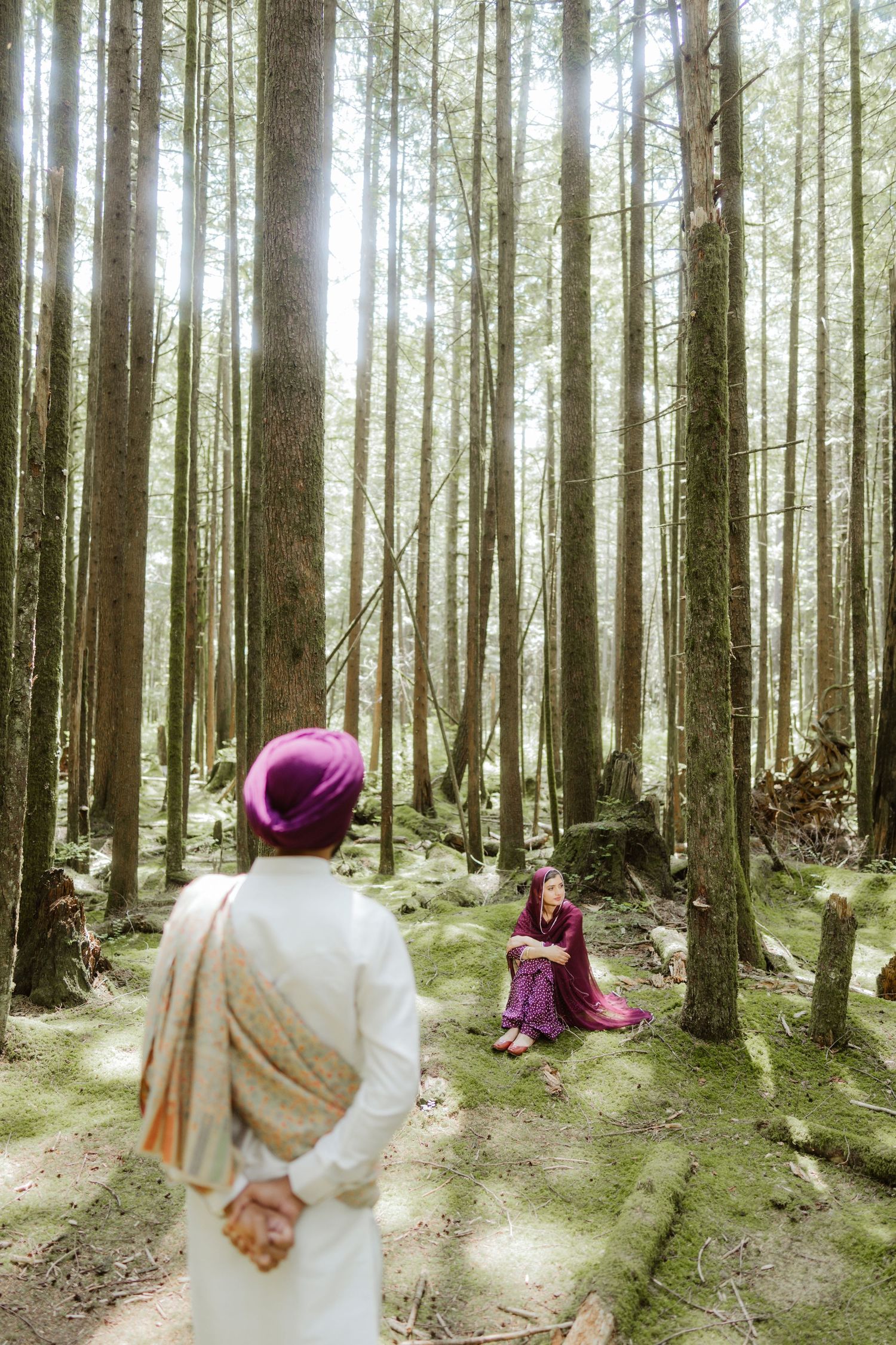 Sikh couple in forest, man in white kurta and purple turban walks toward woman in purple salwar kameez sitting among tall trees.