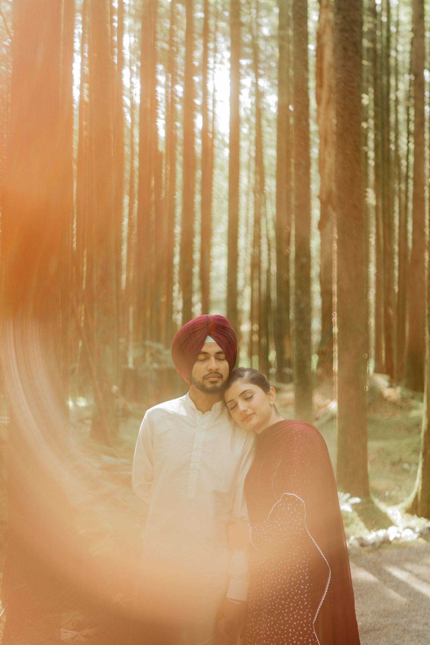 Sikh couple in forest with golden sunlight, man in red turban and white kurta, woman in dark maroon dupatta leaning on his shoulder.