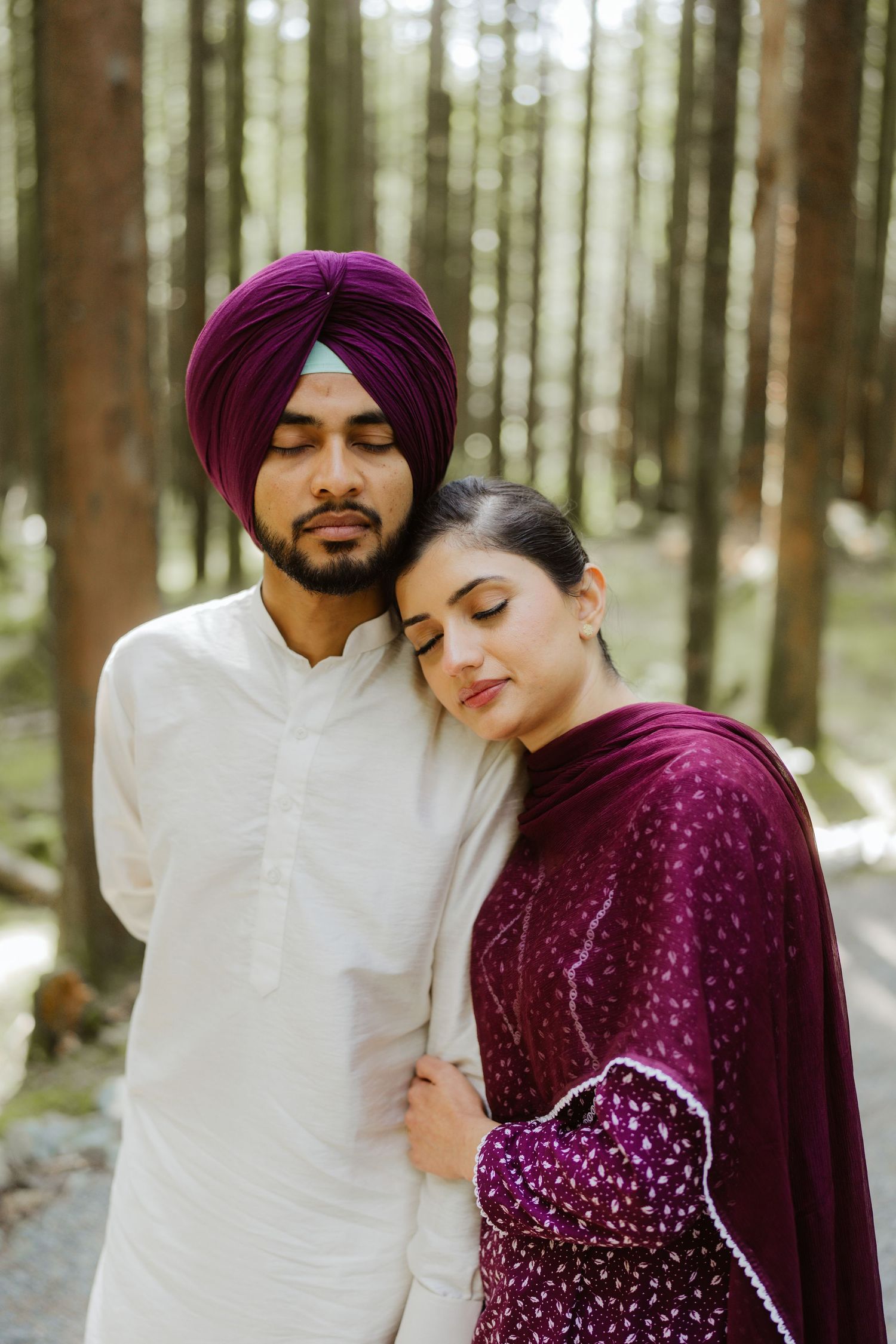 A Sikh couple embraces in a forest, man wearing purple turban and white kurta, woman in maroon salwar.