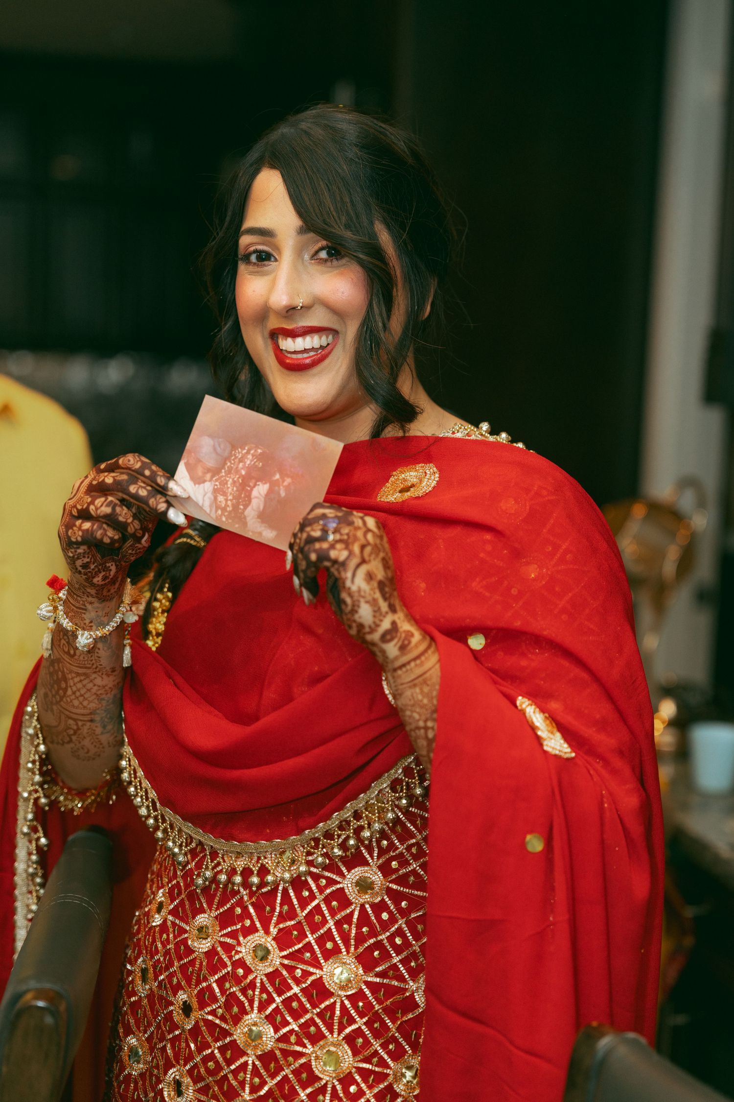 Smiling South Asian bride in red lehenga with gold embroidery and henna hands holding a photo card.