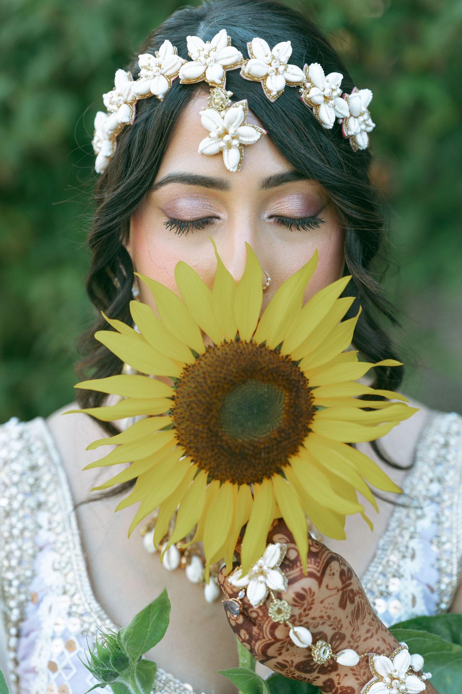 Bride with henna hands holds sunflower near face, wearing floral crown and white dress, eyes closed, outdoors.