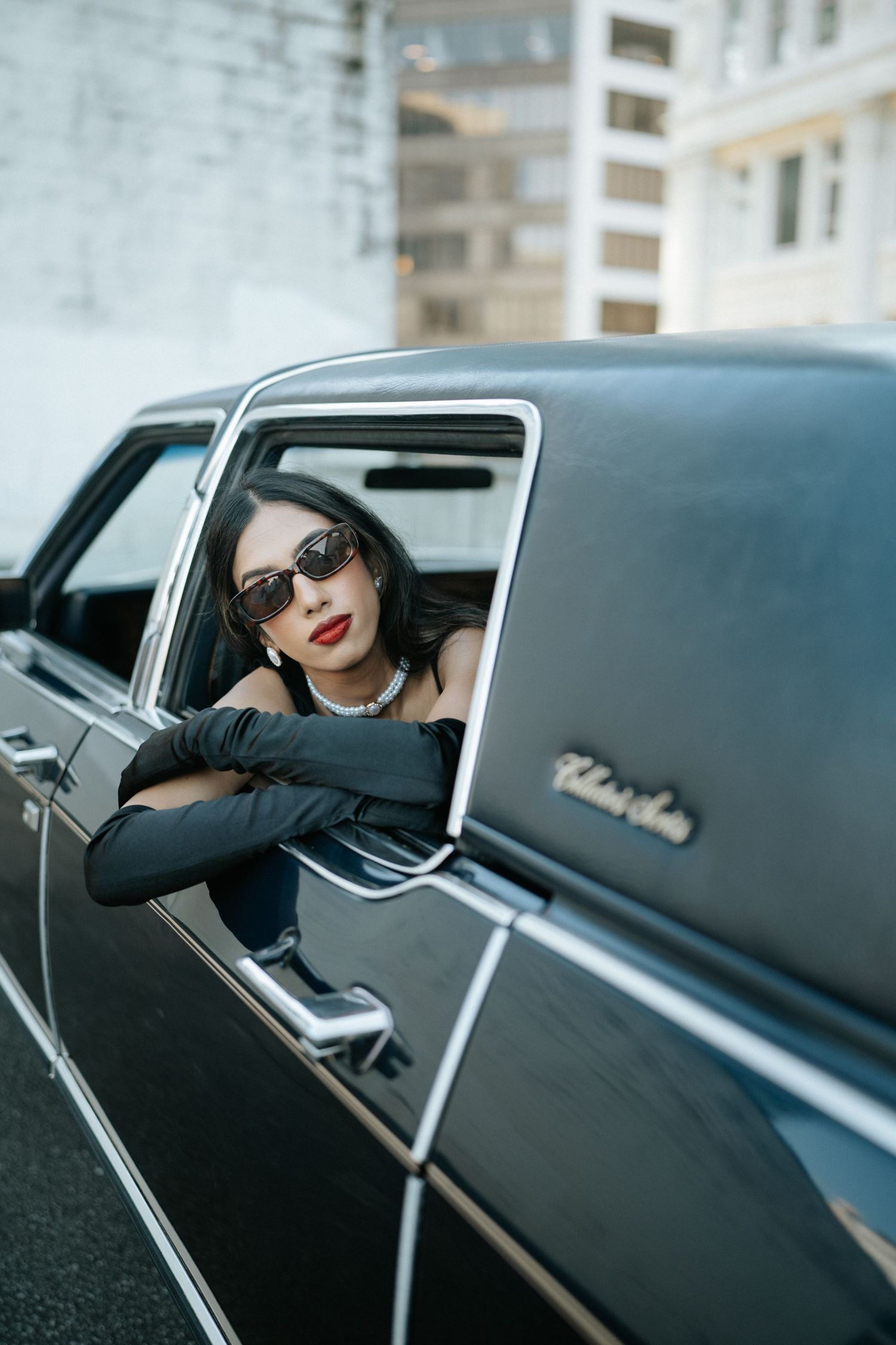 A vintage black car window frames a glamorous figure wearing red lipstick and sunglasses in a retro-style photo series.