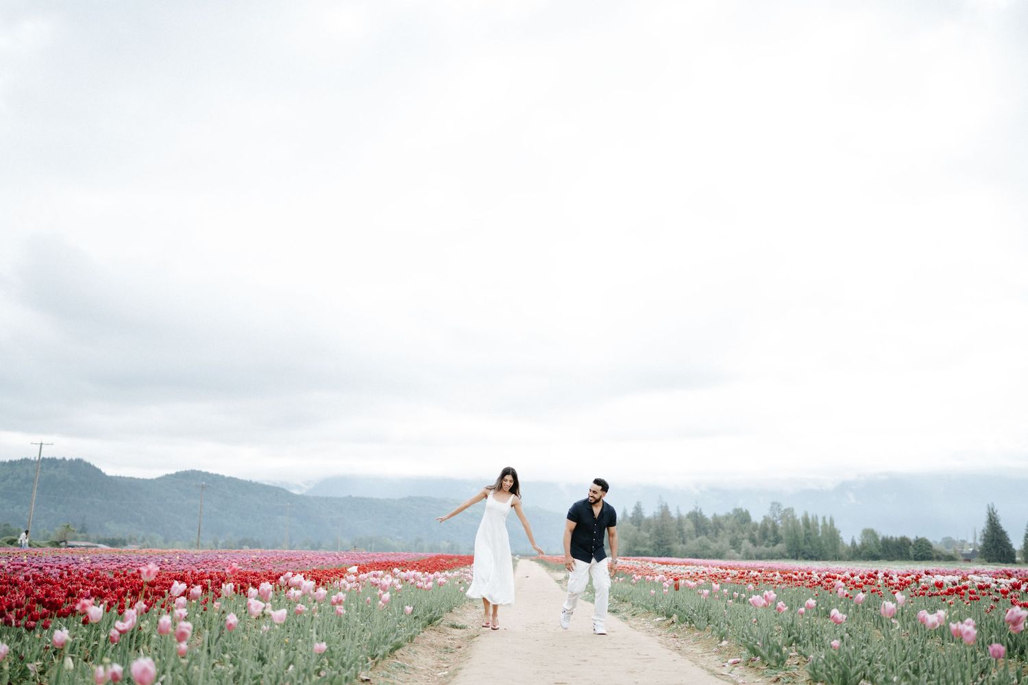 A couple walks through a vibrant tulip field with mountains in the background on a cloudy spring day.