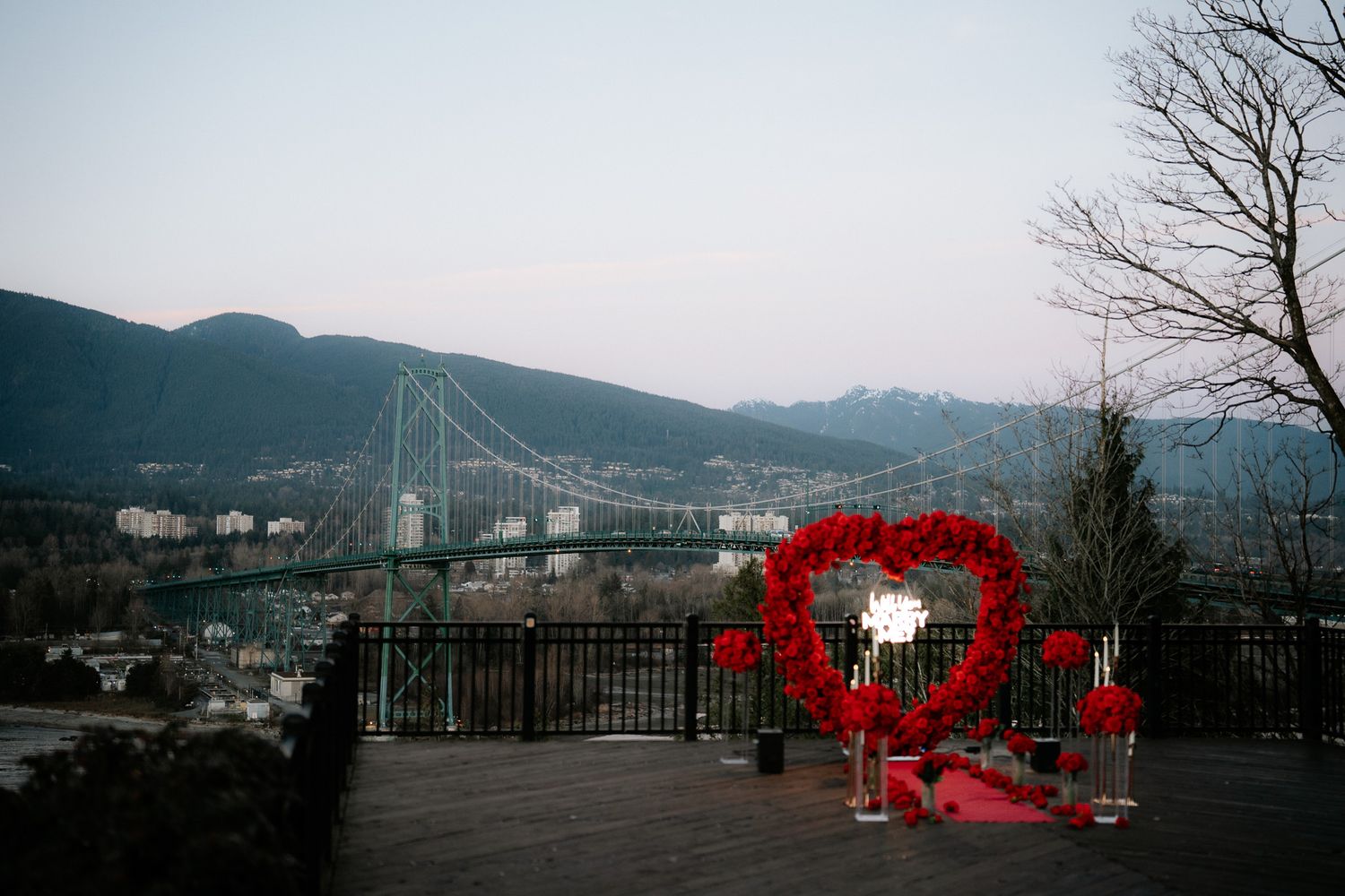 A red heart-shaped art installation overlooks Lions Gate Bridge and mountains in Vancouver at dusk.