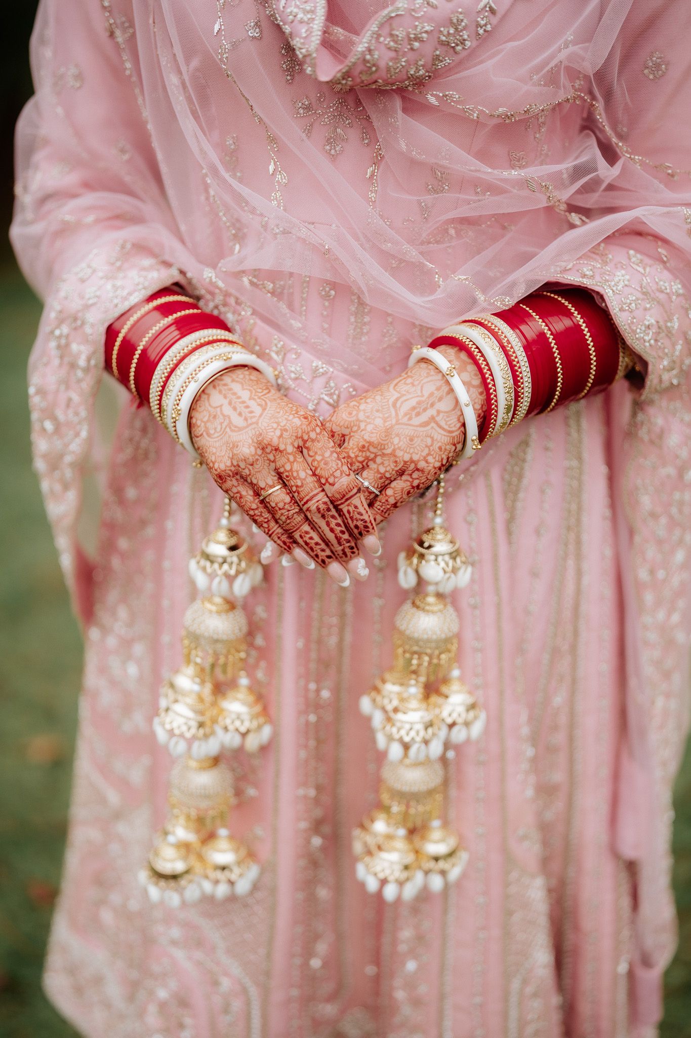 Close-up view of henna-adorned hands holding ornate pearl and gold kalire bridal jewelry against a pink embroidered dress.