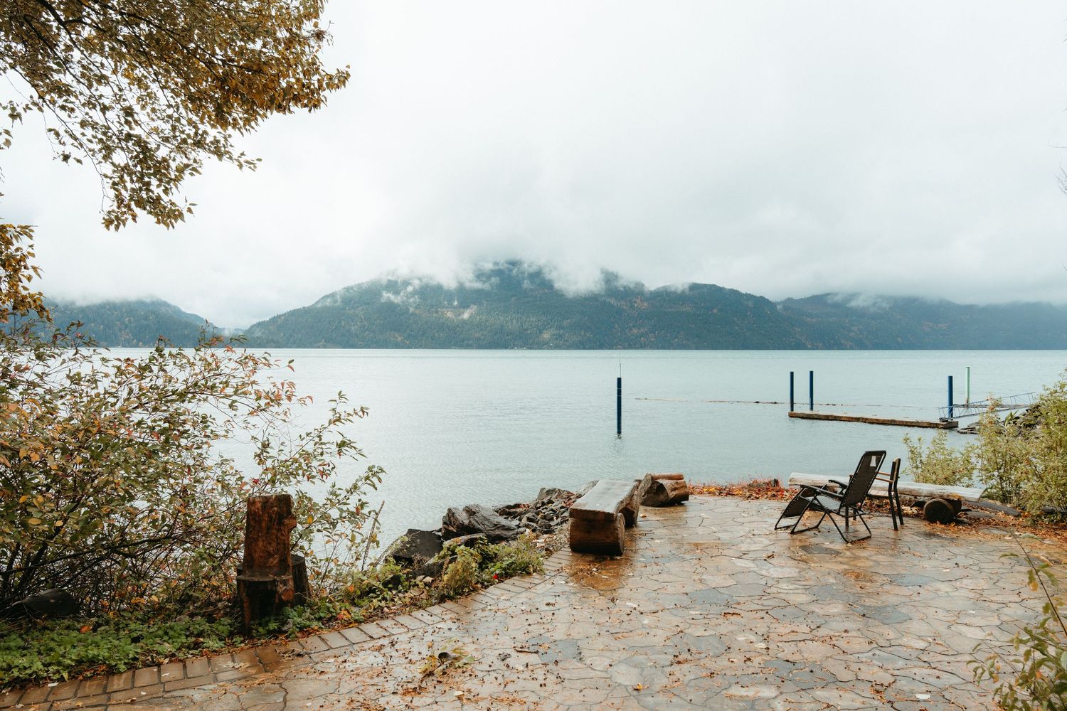 Misty lake view with wooden dock posts and rocky shoreline surrounded by mountains and autumn foliage.