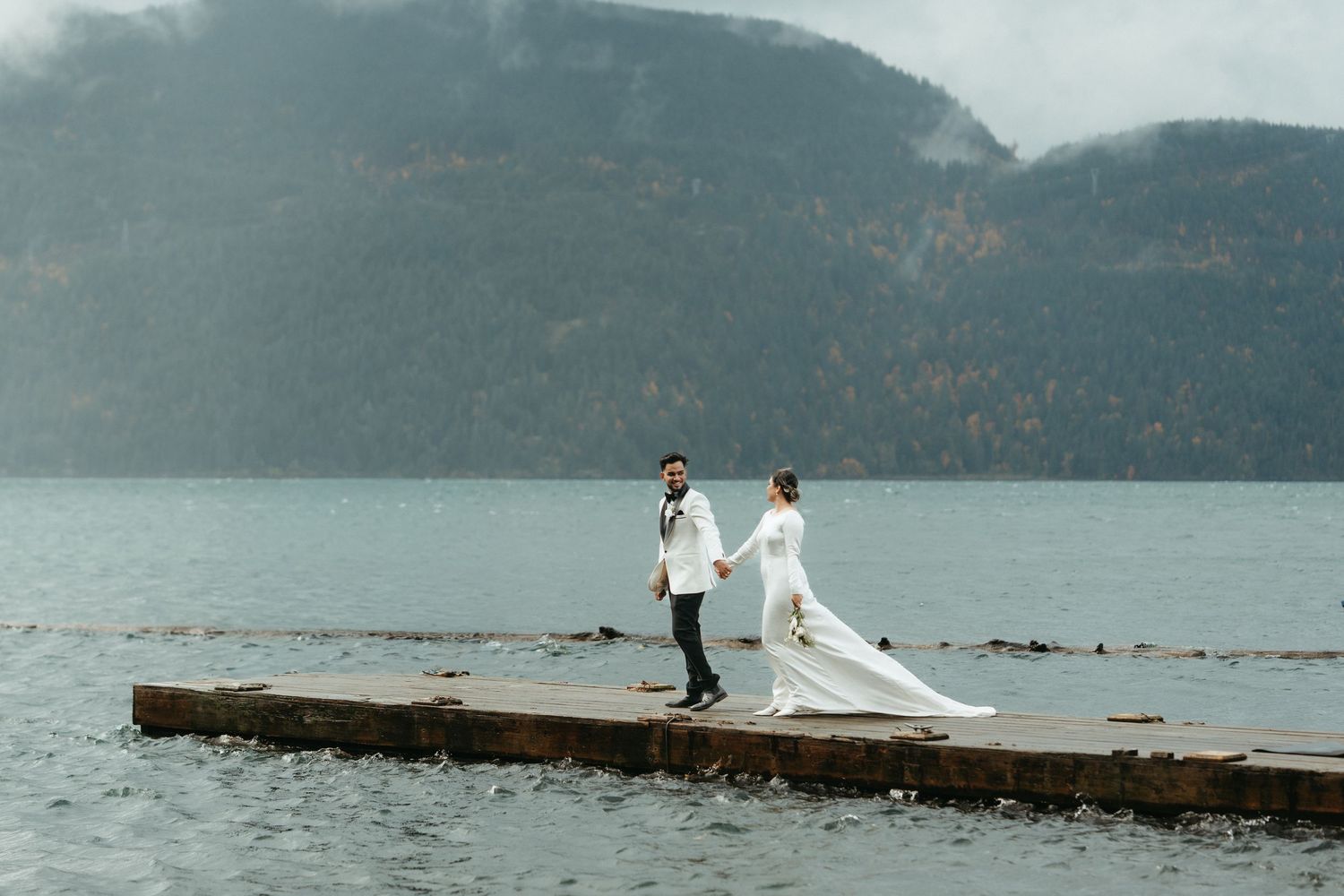 A couple in wedding attire stands on a wooden dock overlooking a misty mountain lake on an overcast day.