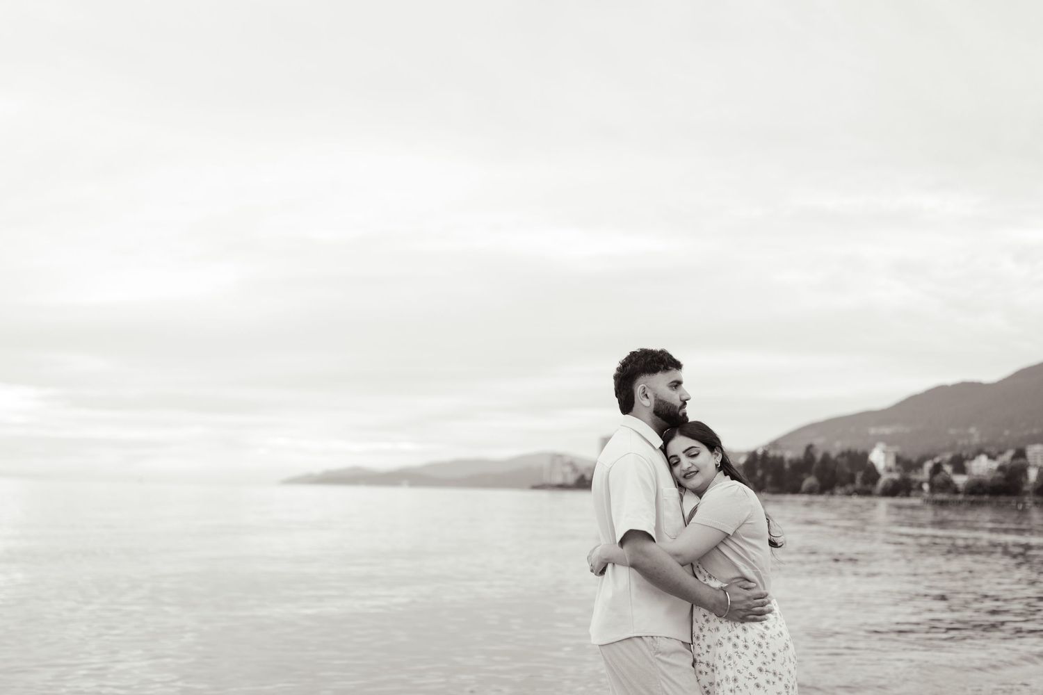 A couple embraces on a peaceful beach with mountains and cityscape in the distance.