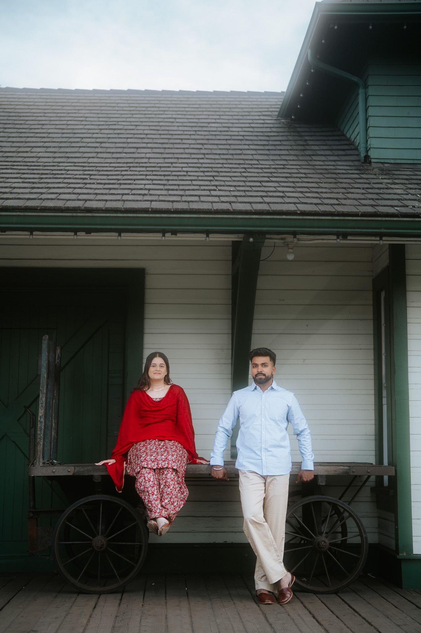 Couple posing on vintage wooden cart outside historic Fort Langley building, woman in red outfit, man in blue shirt.