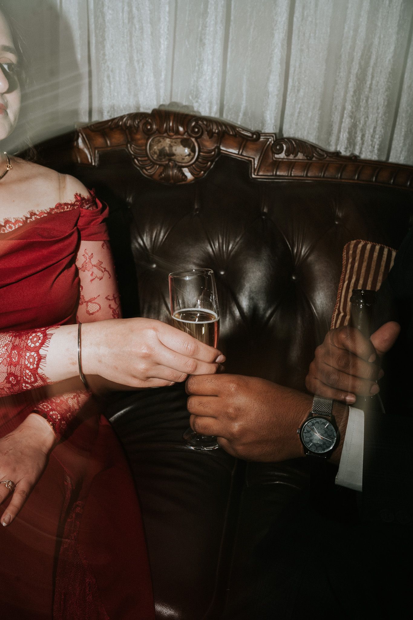 Close-up of couple on dark leather sofa, woman in red lace dress and man in suit sharing champagne glasses.