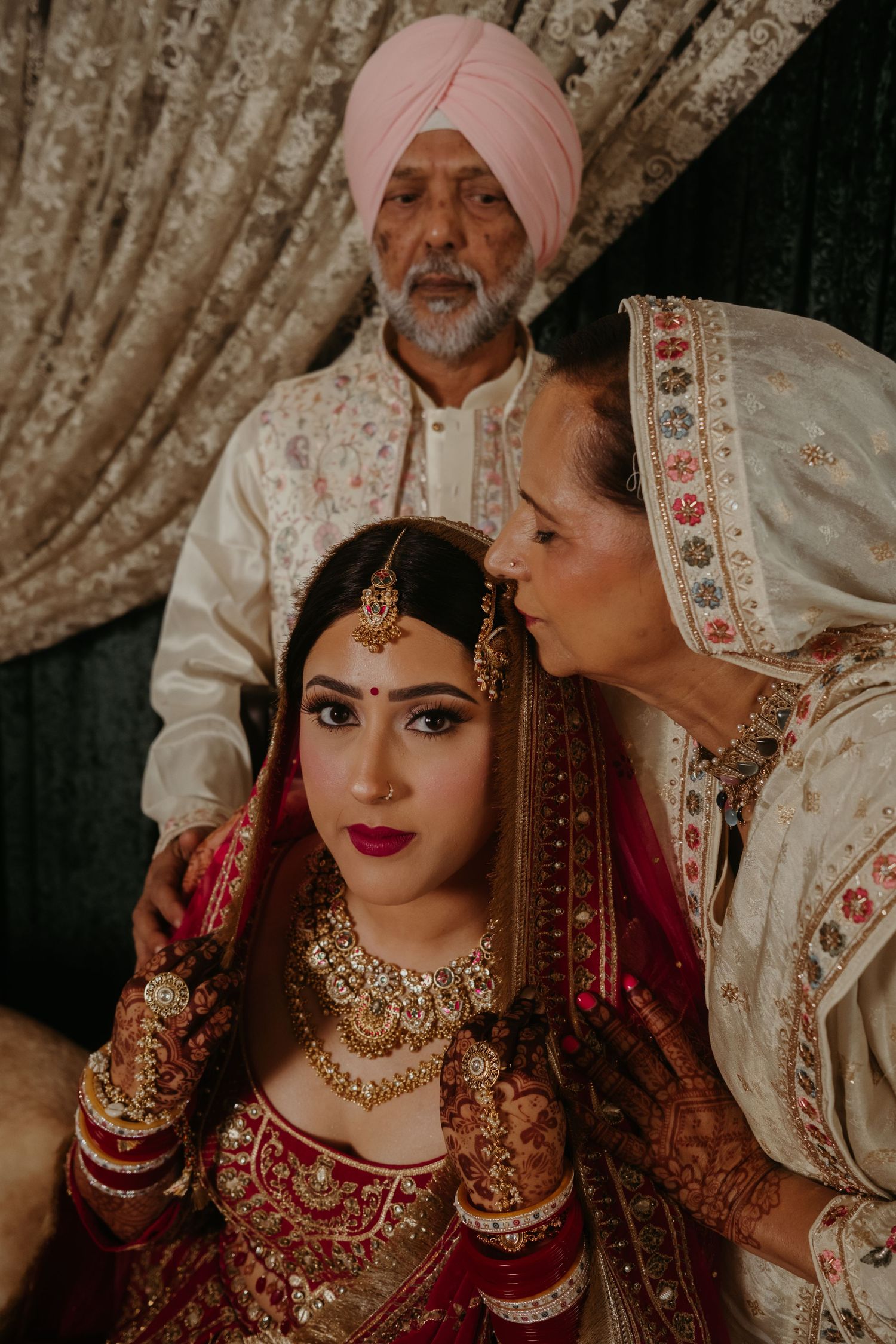 Sikh bride in red lehenga with gold jewelry, flanked by parents in traditional attire during wedding ceremony.
