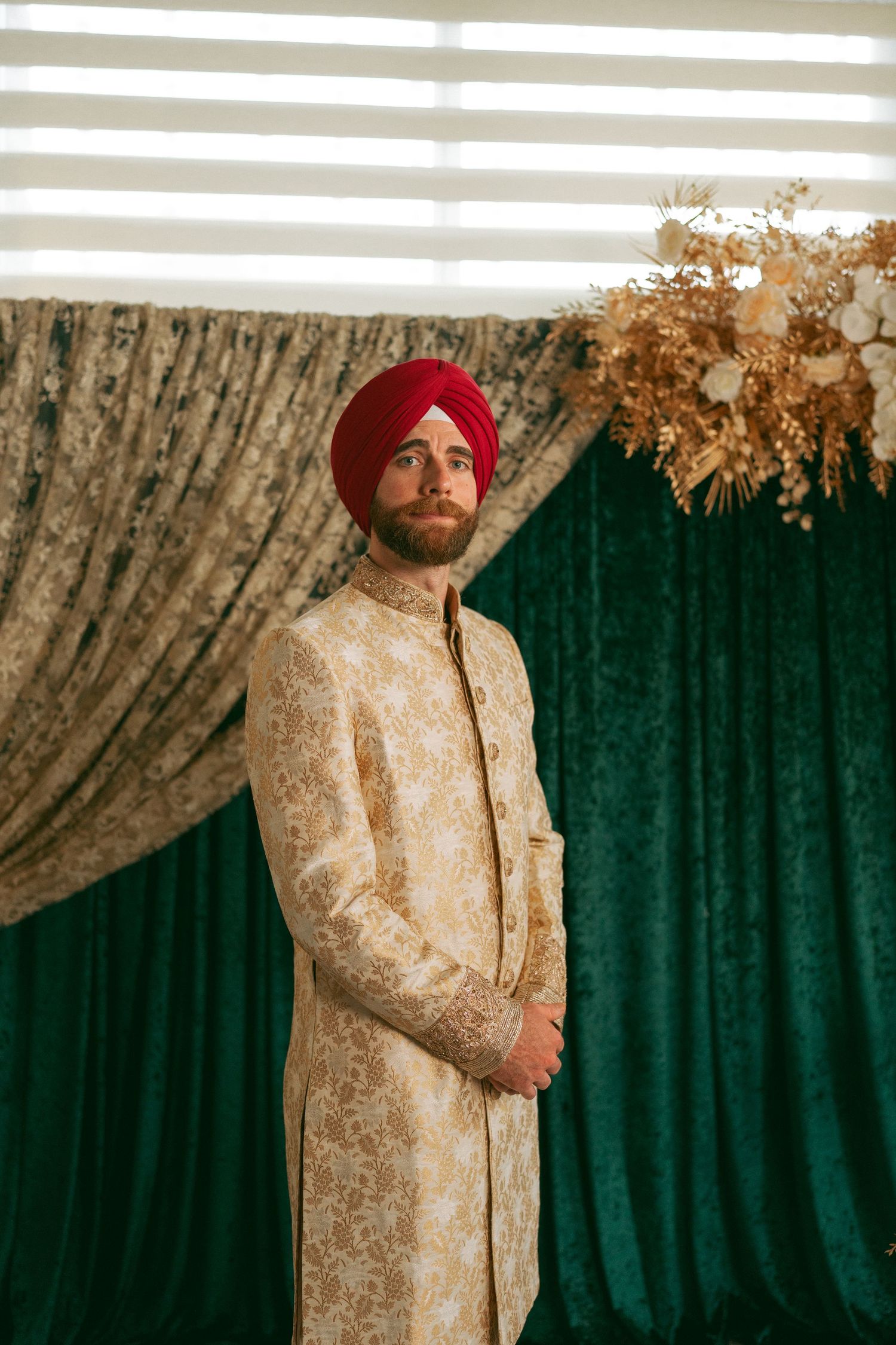 A Sikh groom in a red turban and gold sherwani stands before green velvet drapes and floral decor.