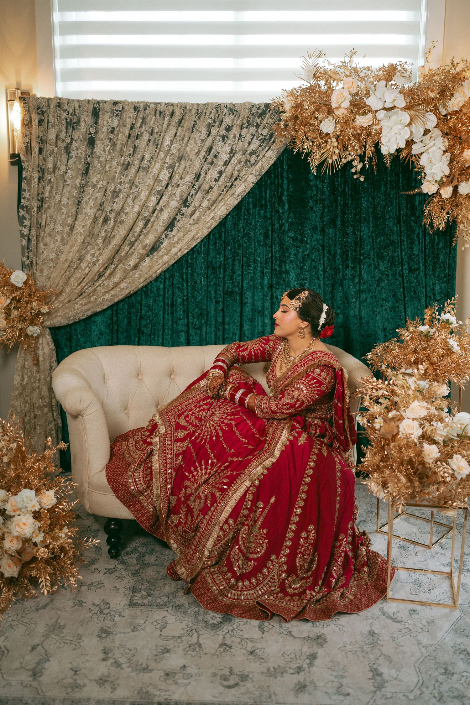 Bride in red and gold embroidered lehenga seated on cream sofa with green velvet backdrop and golden floral decor.