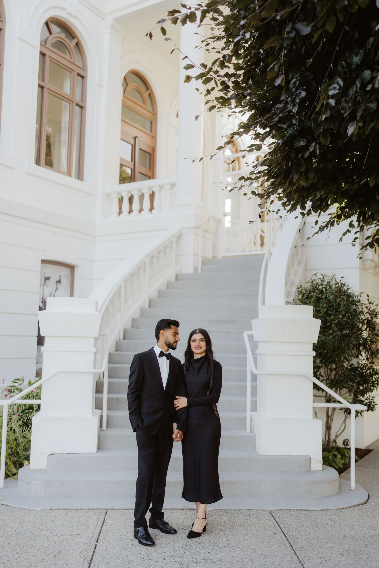 South Asian couple in matching black formal attire standing before a grand white building with elegant staircase.