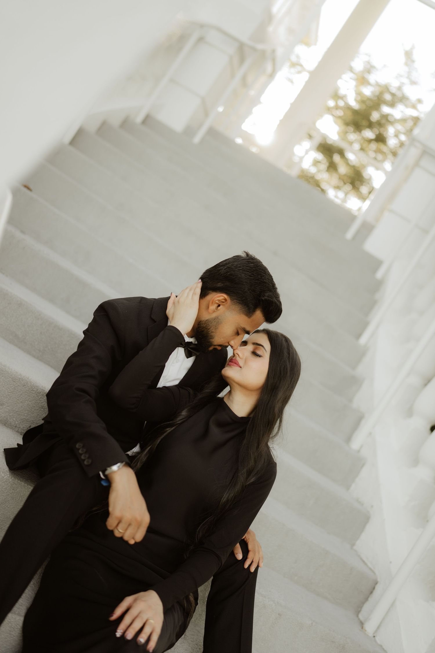 A couple in matching black outfits share a romantic kiss on white marble stairs in an elegant setting.