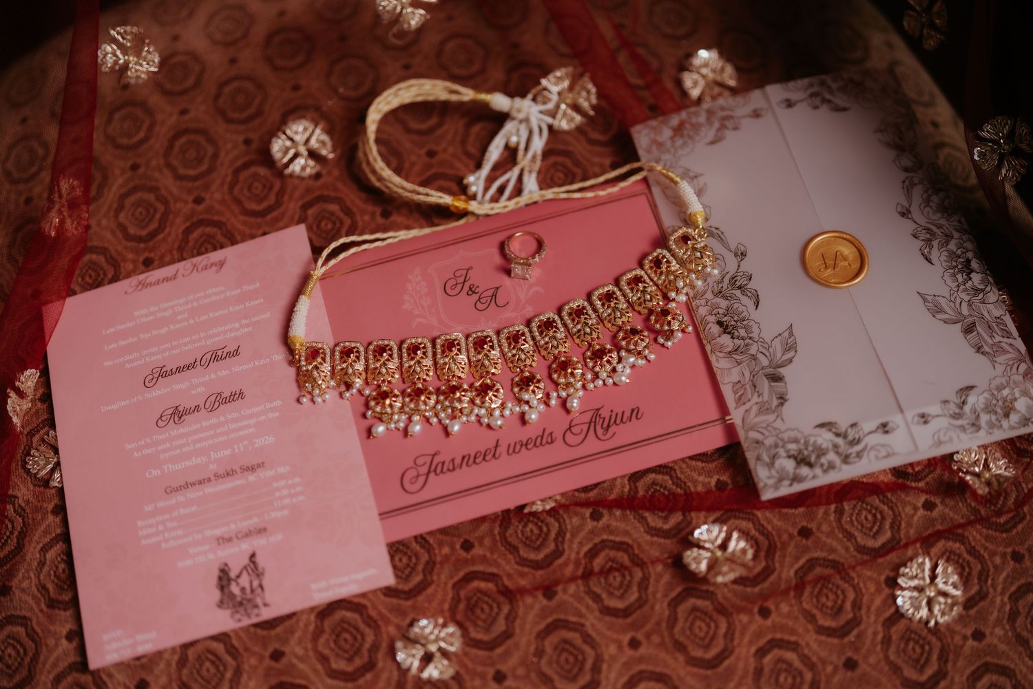 Pink Indian wedding invitation suite with gold necklace jewelry, wax seal envelope, and ornate floral stationery on red fabric.