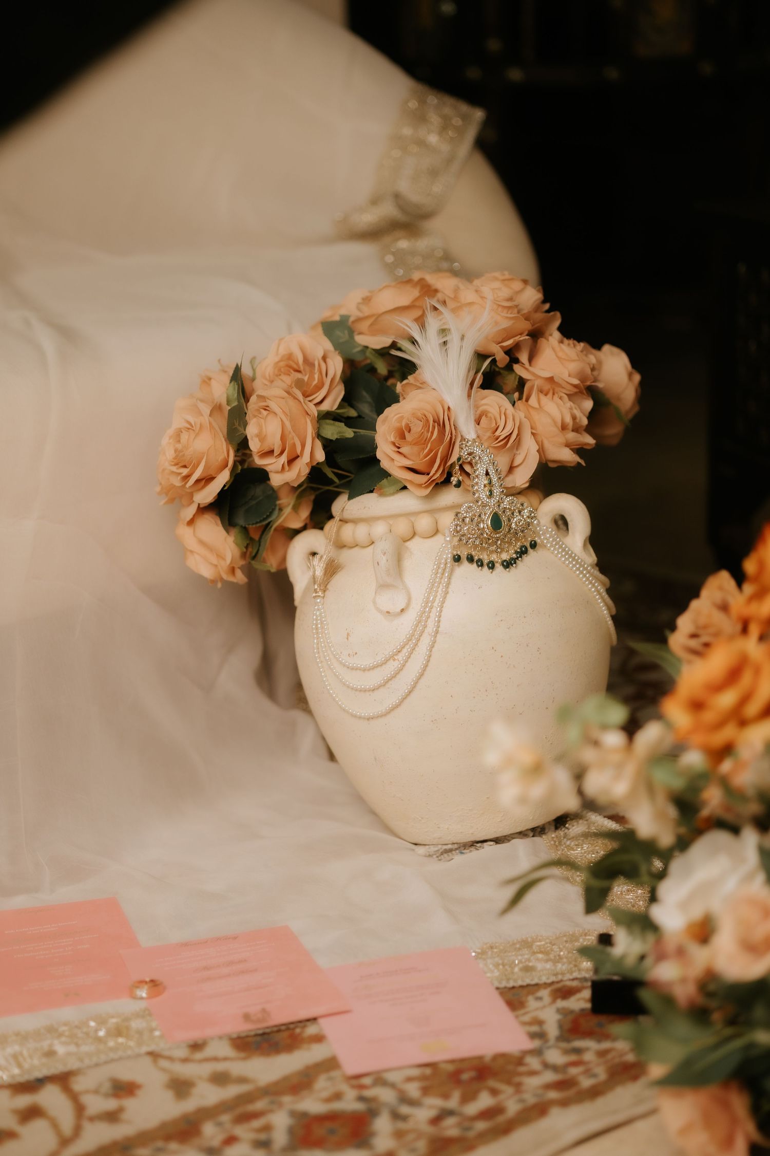 Cream ceramic vase with pearl chains holds peach roses and feather brooch on a decorated wedding table.