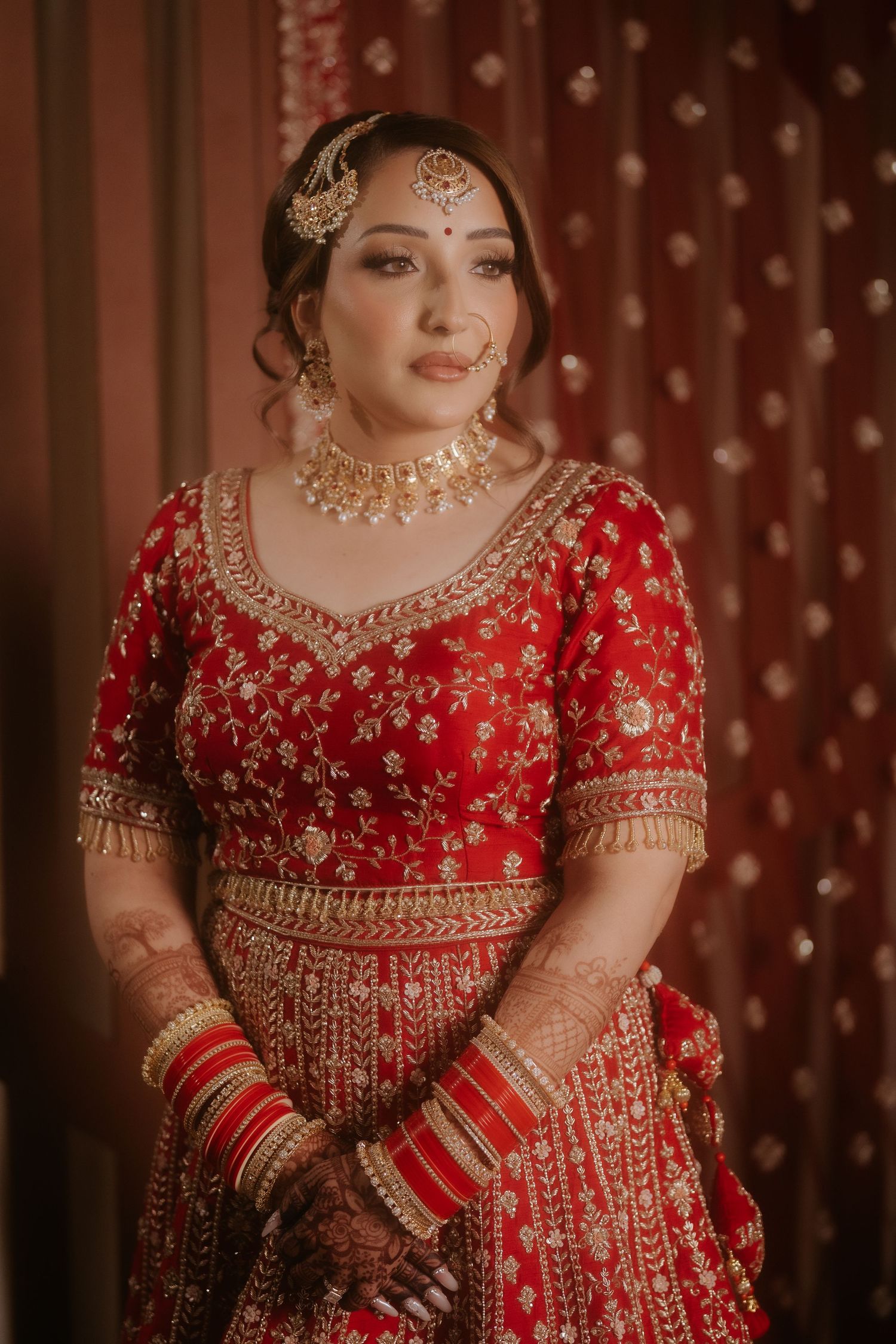 Indian bride in red and gold embroidered lehenga with traditional jewelry, mehndi, and bangles on her wedding day.