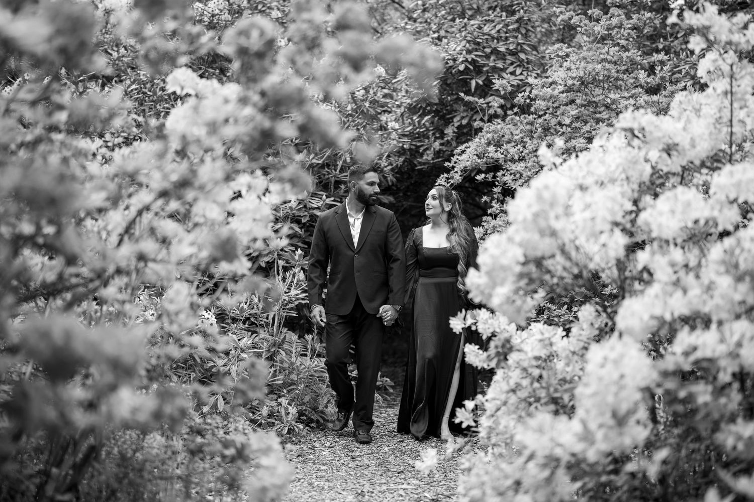 Couple walking hand in hand through lush flowering garden path in elegant black attire, black and white photo.