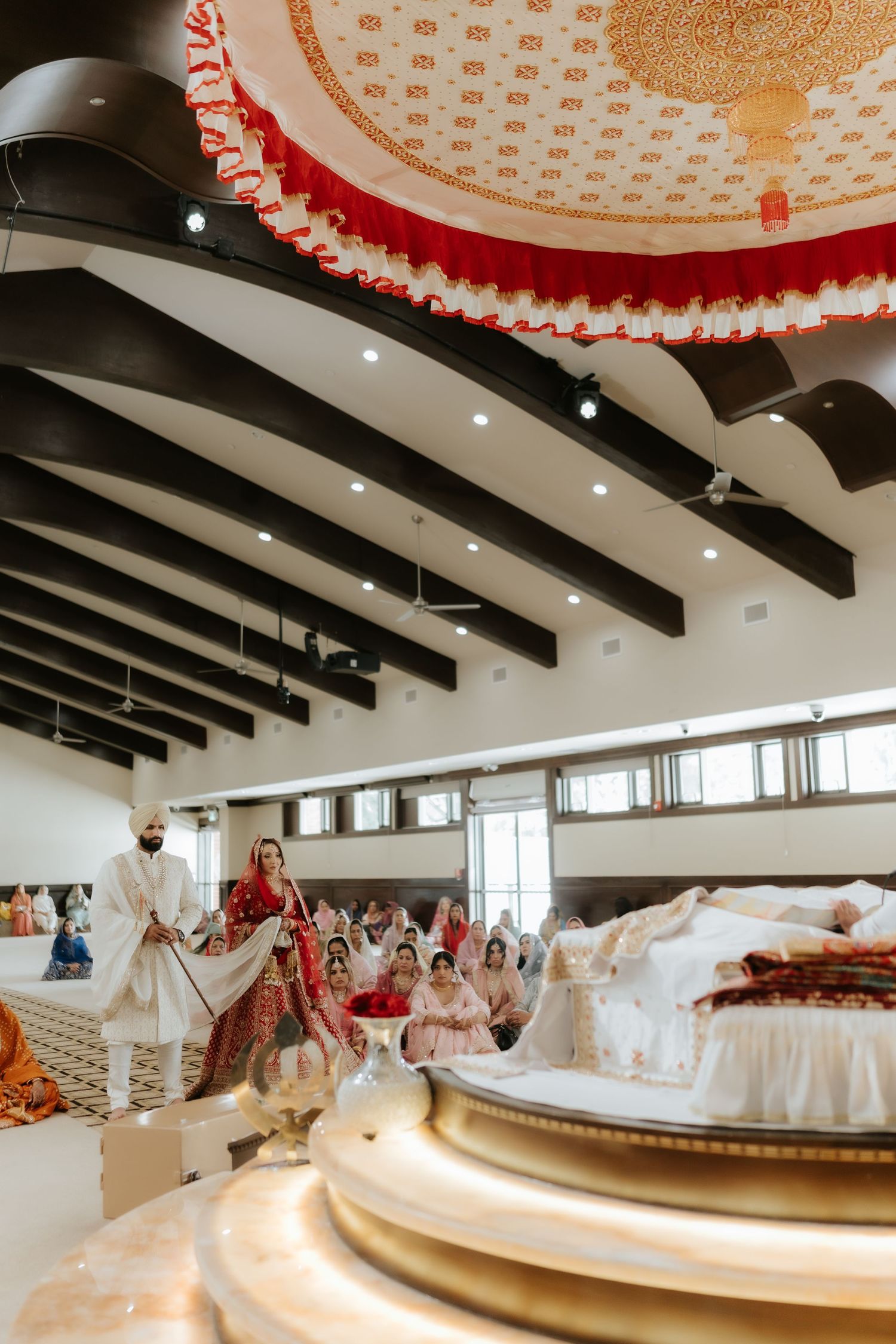 Sikh wedding ceremony in a gurdwara with bride and groom in traditional red and white attire walking around the Guru Granth Sahib.